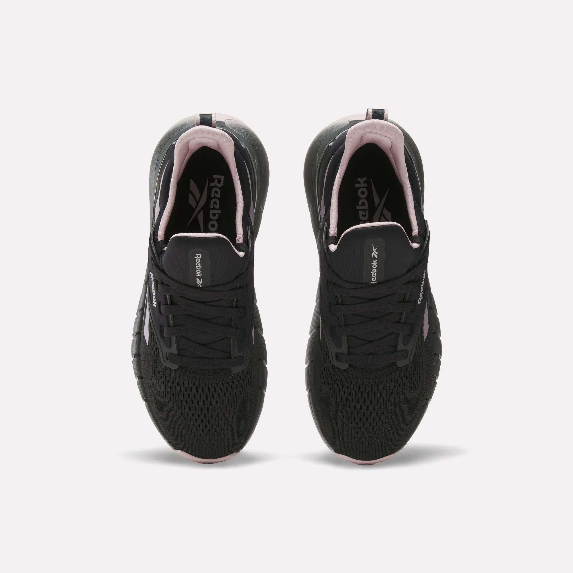 A pair of Reebok Women's Nano Gym Training Shoes in black with white soles and pink accents, shown from above on a white background. Versatile for workouts, they feature the Reebok logo on the insoles.
