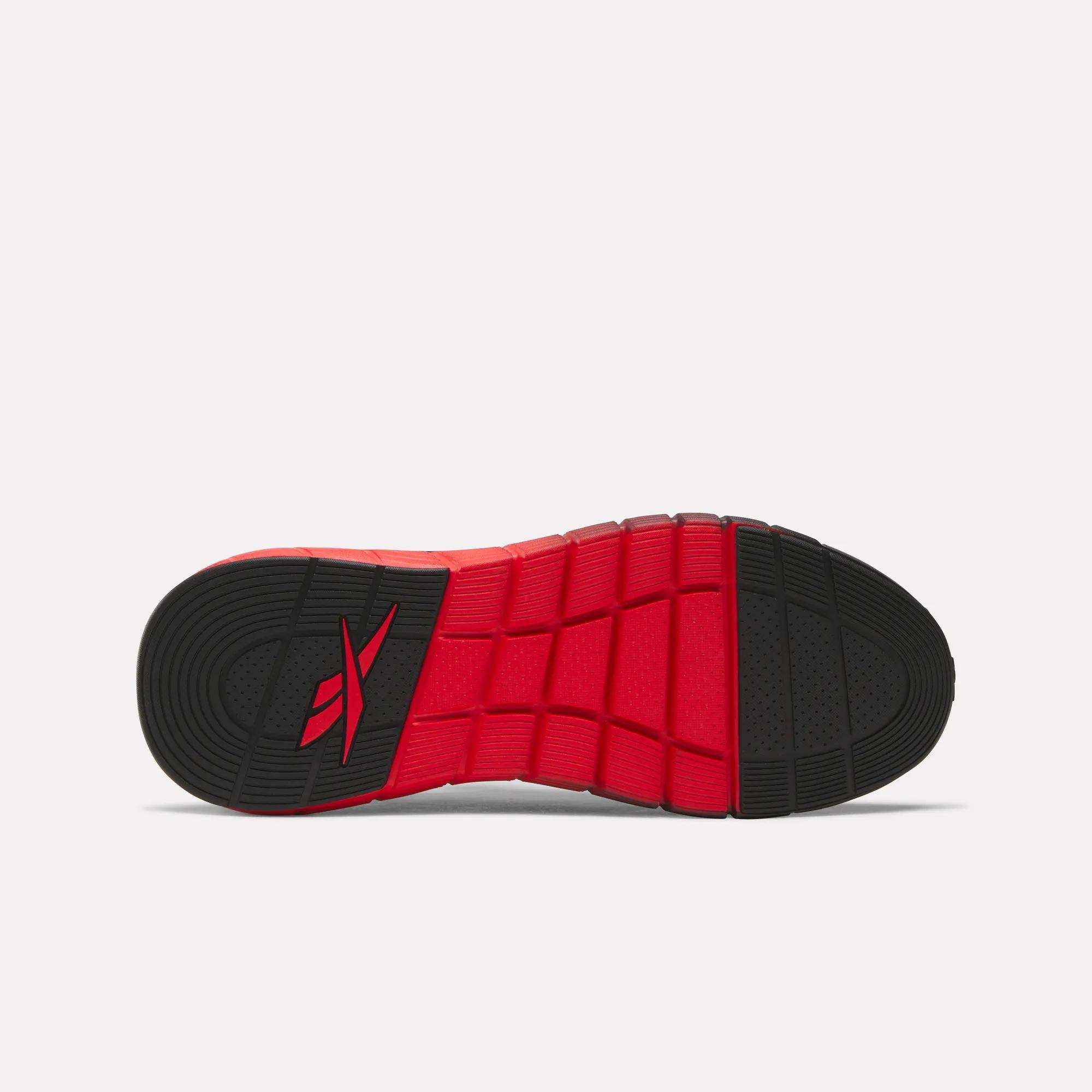 The image displays the sole of Reebok Men's Nano Gym Training Shoes, featuring a ribbed texture with a red and black color pattern. The red Reebok logo appears near the heel on the black section, highlighting its distinctive training shoe style.