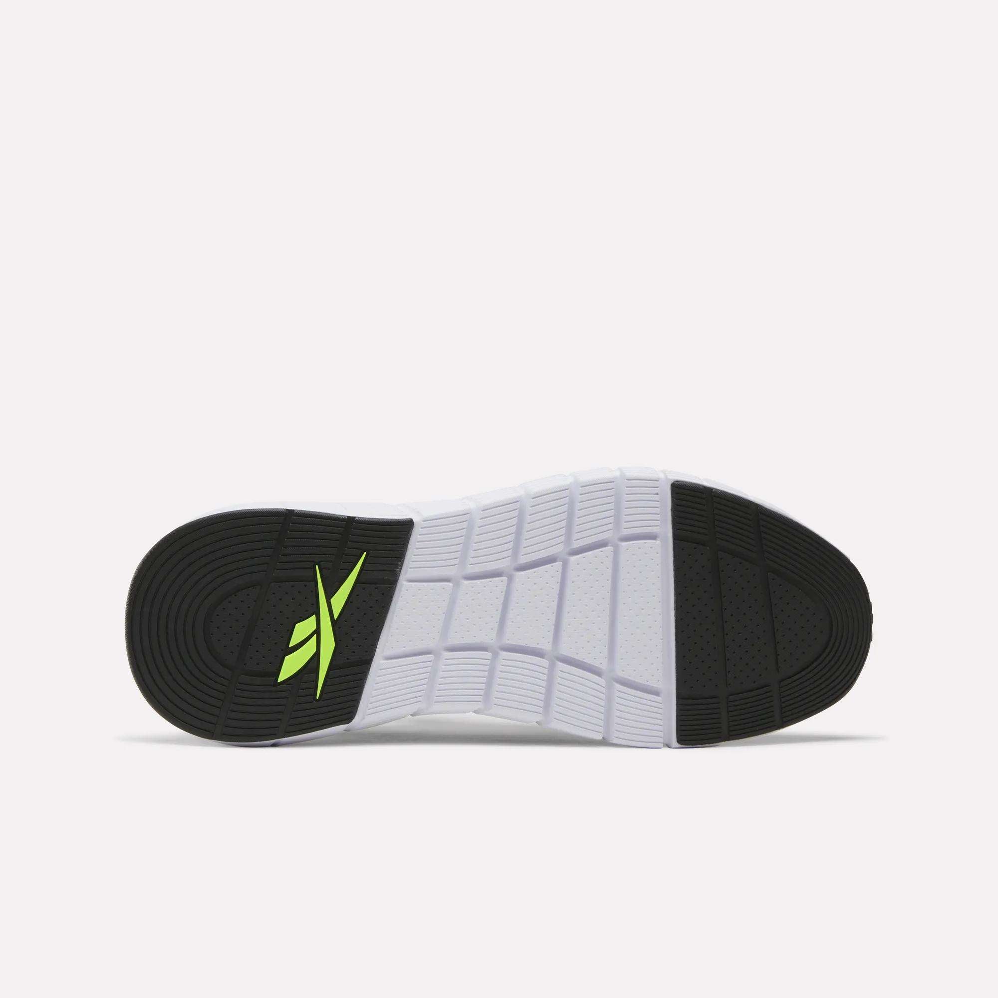The image shows the sole of a white Reebok Men's Nano Gym Training Shoe with black heel and toe sections, a yellow-green Reebok logo near the heel, and a textured grid pattern—ideal for versatile workouts.