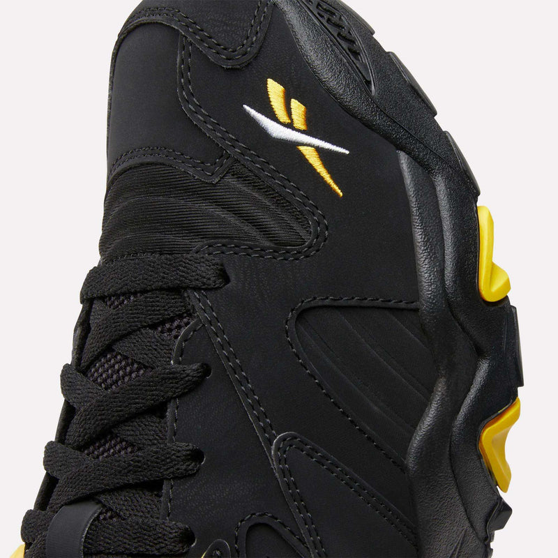 Close-up of a black Reebok Brutus Low Shoe with black laces, yellow and white stitched Reebok logo, and yellow accents on the side of the sole. Detailed stitching and textured materials offer authentic street-ready style.