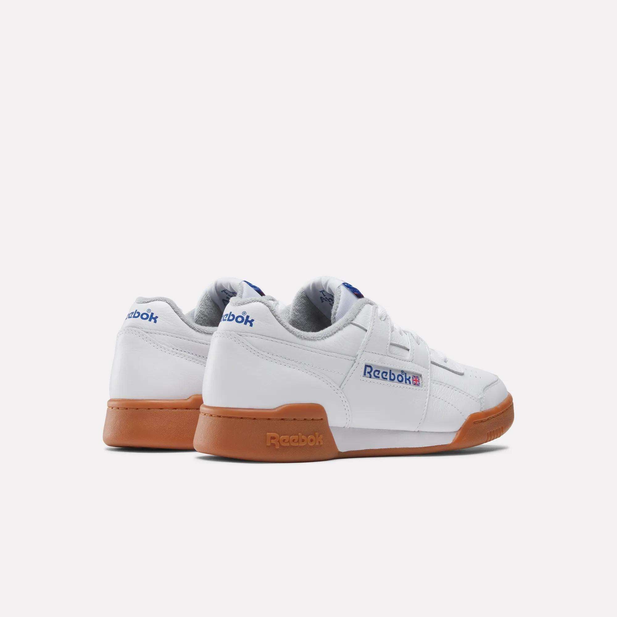 The Reebok Women's Workout Plus Shoes feature a white design with blue accents and gum brown soles, shown angled to display both back and side views. These classic '80s fitness sneakers stand out against a plain white background.