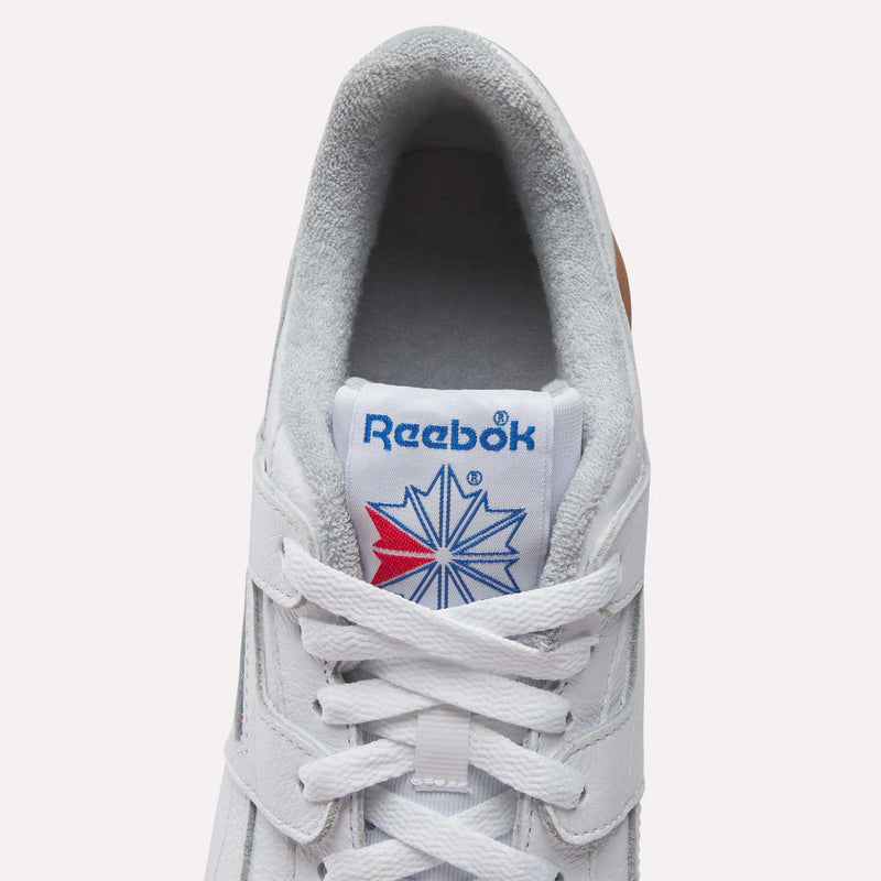 Top view of Reebok Women's Workout Plus Shoes in white, featuring white laces, grey lining, and a blue Reebok logo with red and blue starburst on the tongue—classic '80s fitness style.