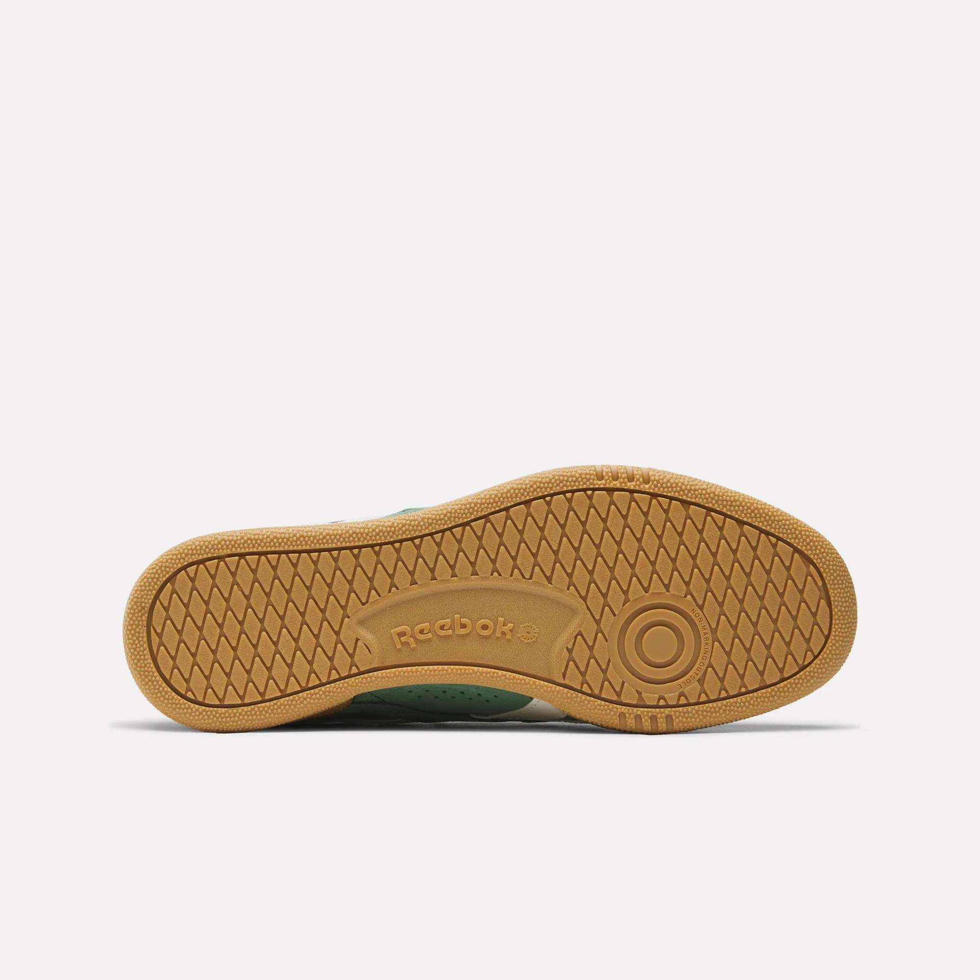 The image shows the bottom view of the Reebok Club C Grounds UK Shoes, featuring a classic design with a textured light brown rubber sole, circular pattern near the heel, and an embossed Reebok logo at the center.
