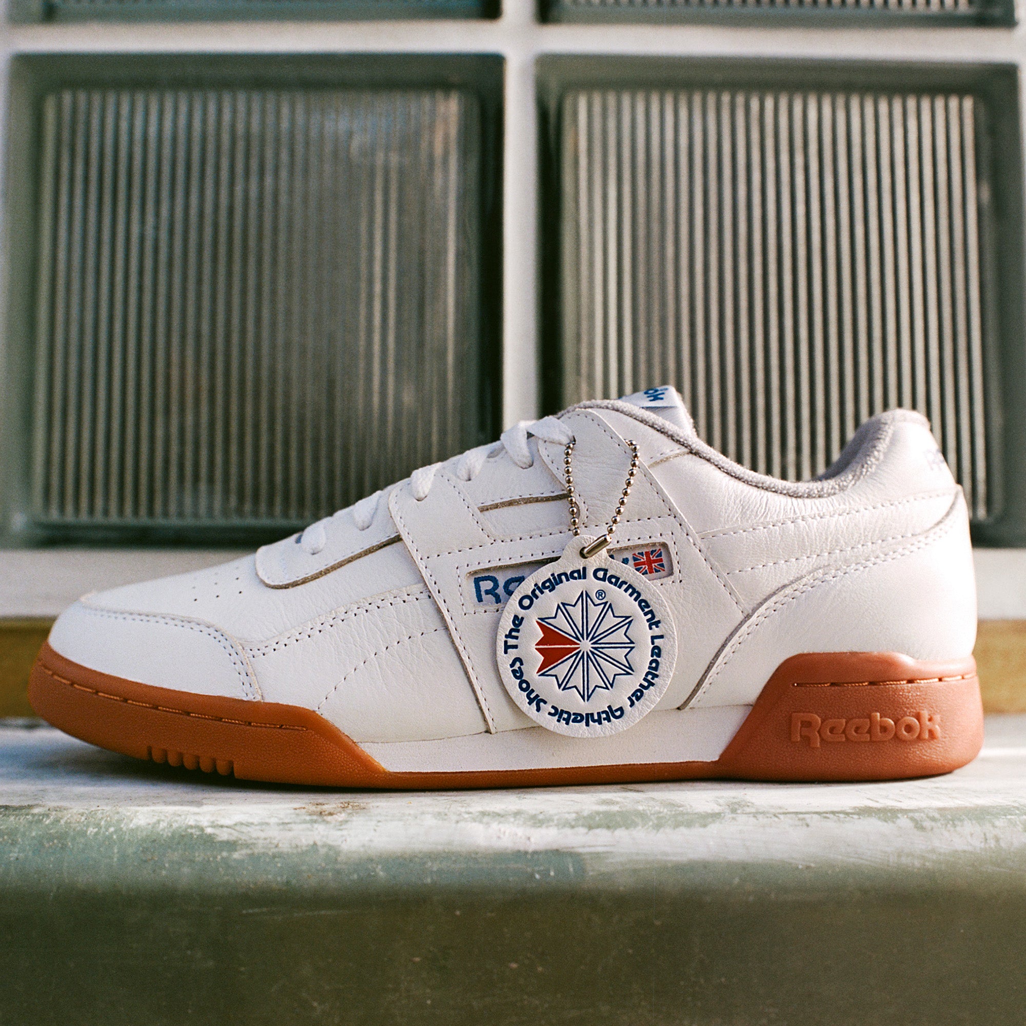 The Reebok Workout Plus Shoes feature a white upper, gum sole, blue accents, and a round tag reading "The Original Garment Leather Reebok," capturing '80s fitness style against frosted glass tiles.