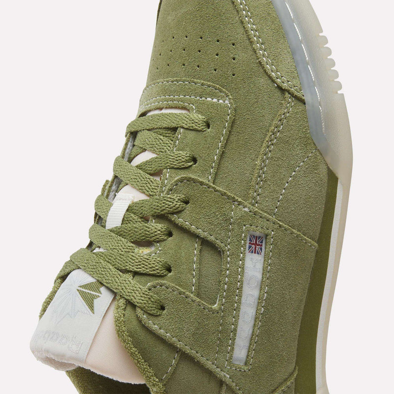 Close-up of olive green suede Reebok Workout Plus Shoes with beige laces, white accents, and the iconic logo and Union Jack on the side. Detailed stitching and a textured rubber sole evoke classic '80s fitness style.