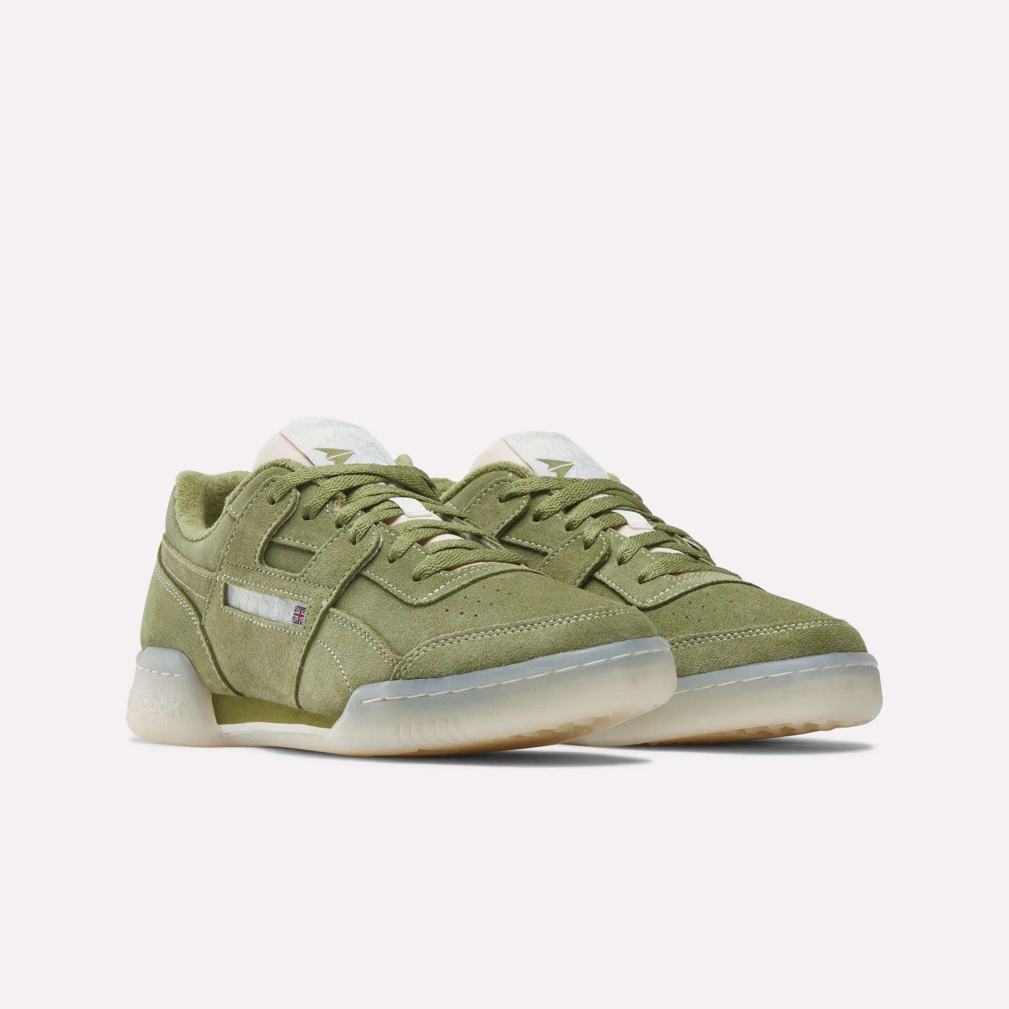 The Reebok Workout Plus Shoes are olive green low-top sneakers with suede uppers, white tongues, and translucent rubber soles, inspired by '80s fitness style. Displayed on a plain white background.