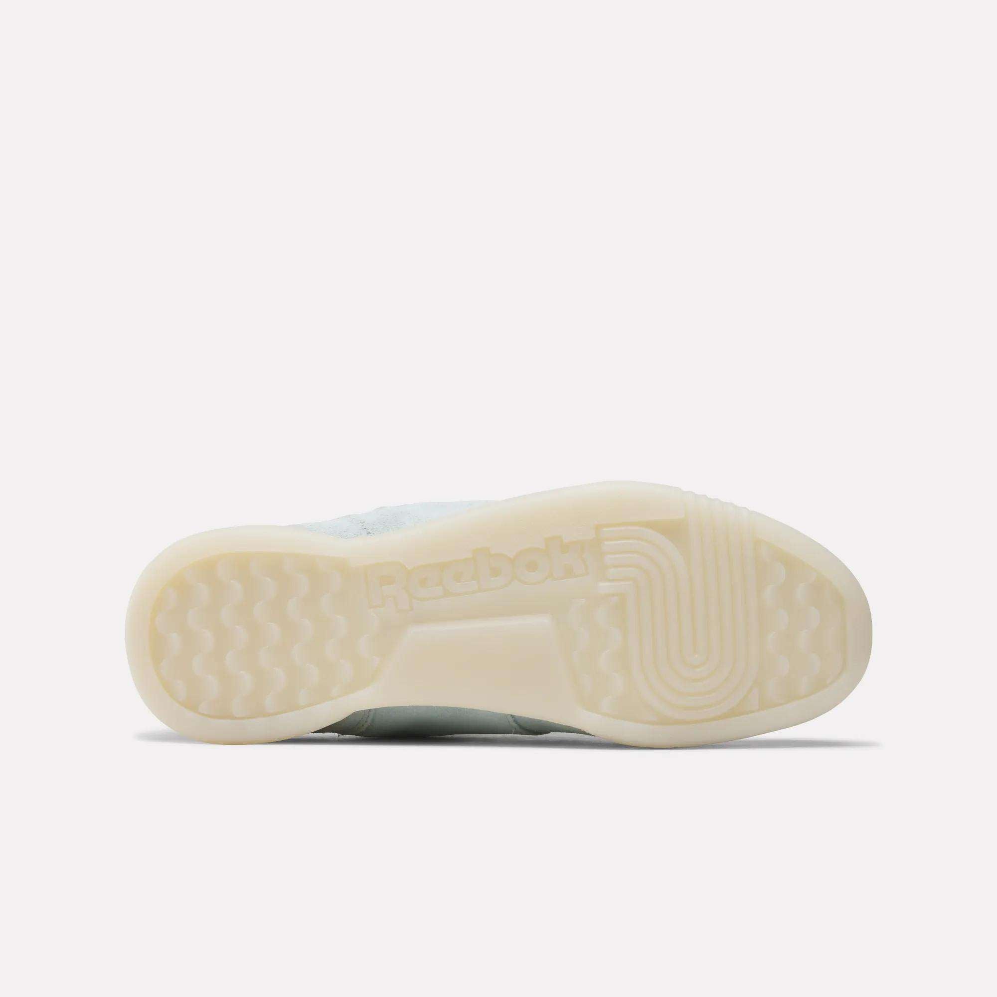 The image displays the bottom view of Reebok Women's Workout Plus Shoes with a translucent rubber sole, classic textured grip patterns, and the Reebok logo—reflecting iconic 80s fitness-inspired design.