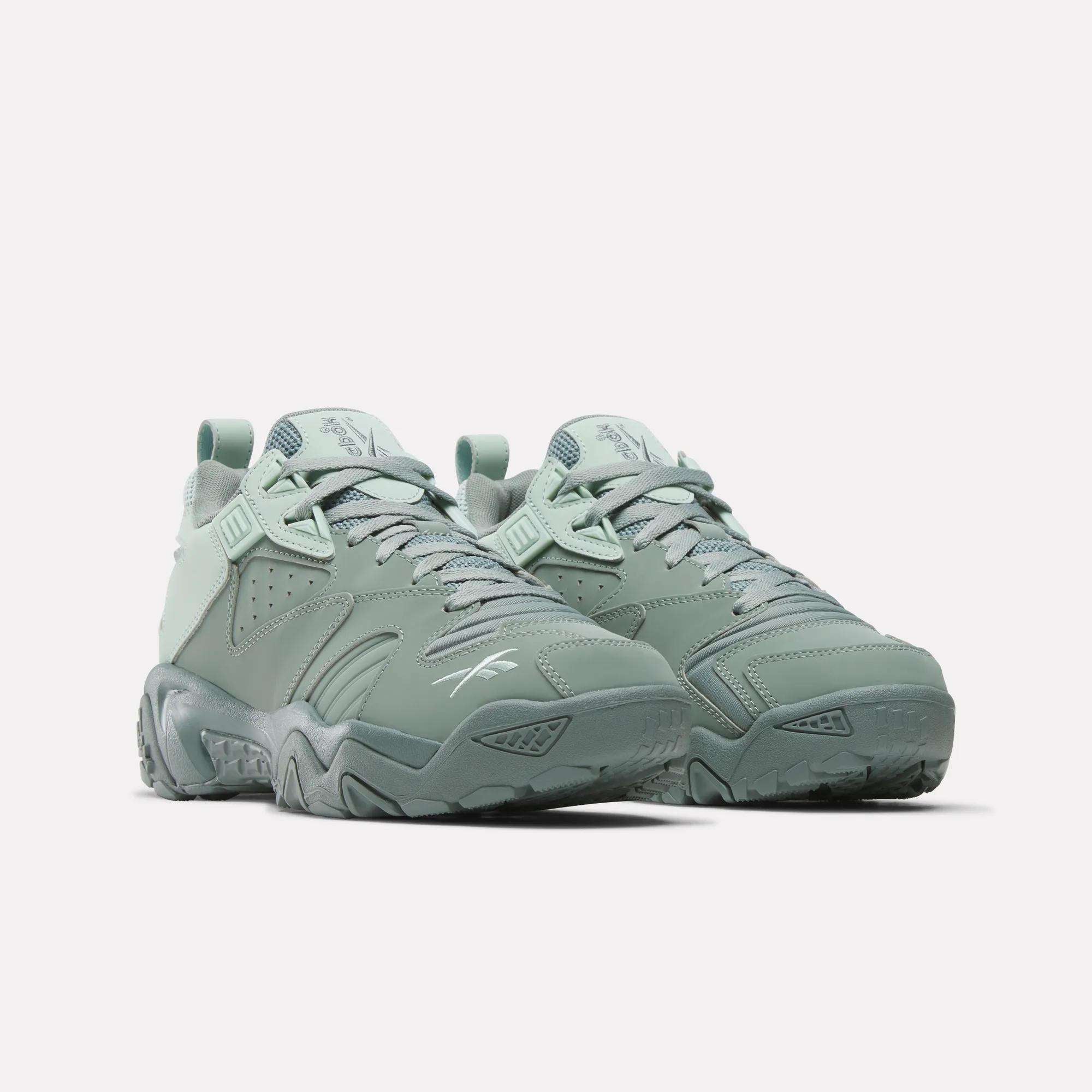 A pair of chunky, sage green Reebok Brutus Low Shoes with lace-up fronts, textured soles, and side Reebok logo. These heritage shoes feature an EVA midsole for added comfort and are shown against a plain white background.