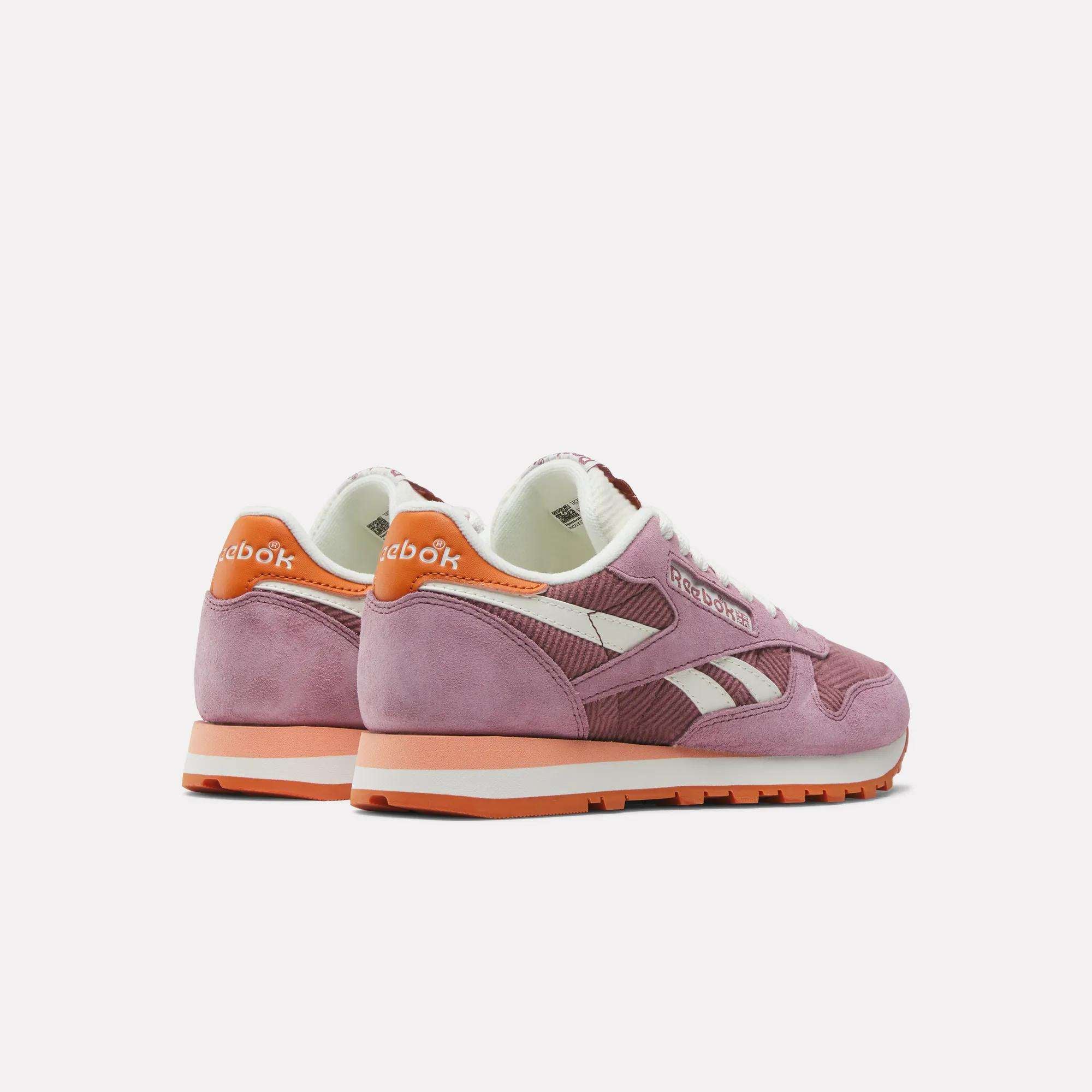 Two Women's Classic Leather Shoes by Reebok in pink, featuring white laces, suede and corduroy details, and orange heel tabs, are shown on a plain white background, with both the side and back of the shoes visible.