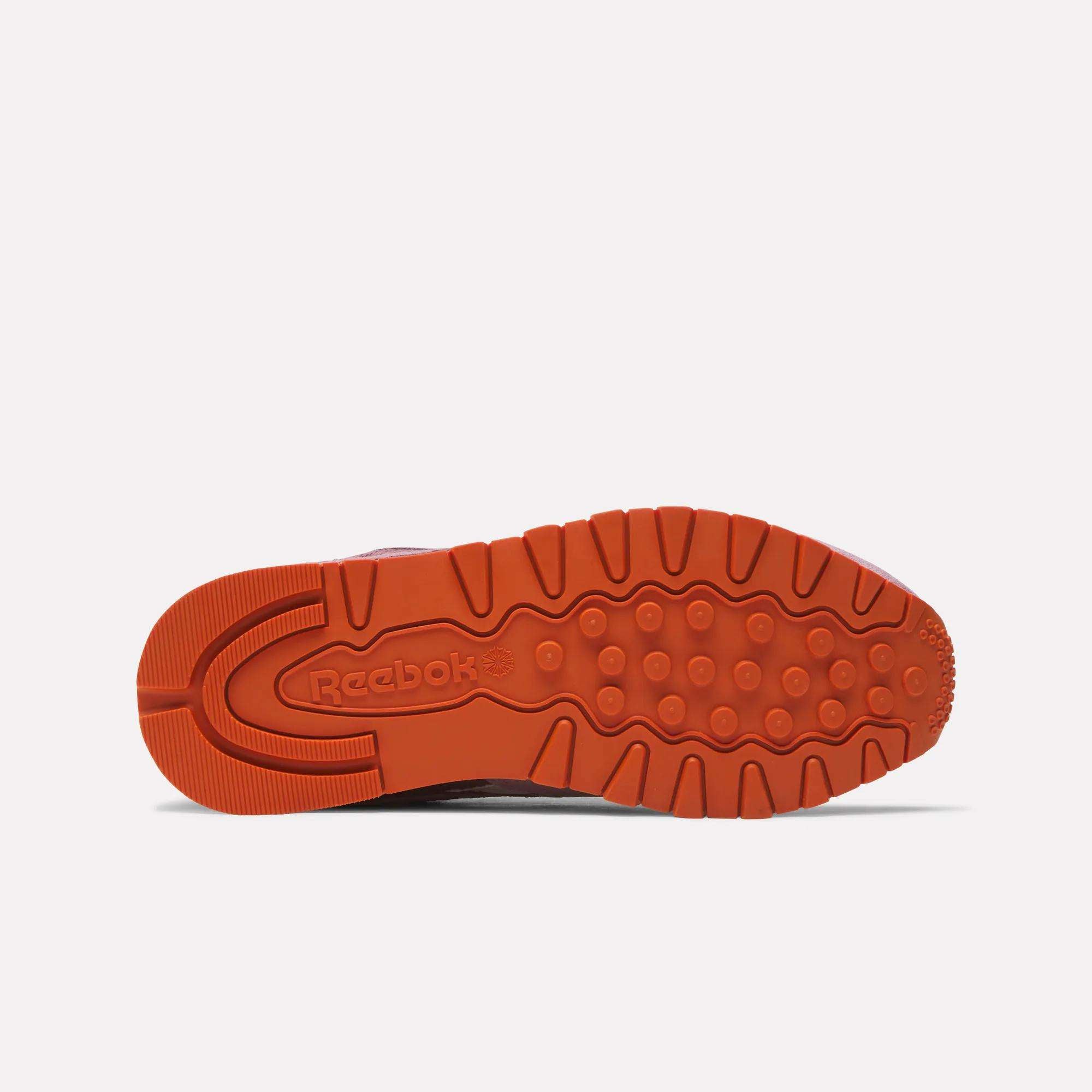 The image shows the orange rubber sole of the Reebok Women's Classic Leather Shoes from below, highlighting various tread patterns and the embossed Reebok logo near the heel.