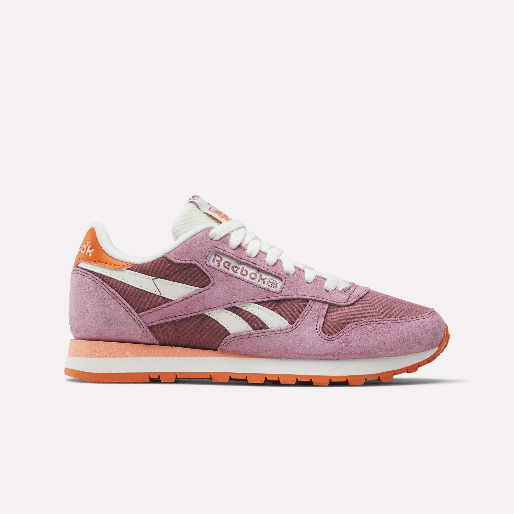 The Reebok Women's Classic Leather Shoes in pink and purple, with white laces, corduroy and suede accents, plus a white sole with orange details, are shown in side profile against a light background.