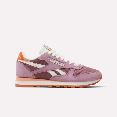 The Reebok Women's Classic Leather Shoes in pink and purple, with white laces, corduroy and suede accents, plus a white sole with orange details, are shown in side profile against a light background.