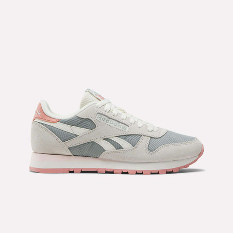 The Reebok Women's Classic Leather Shoes feature light grey and white panels, pale pink accents on the heel, outsole, and branding, suede details, a white lace-up closure, and a flat rubber sole for a retro look.