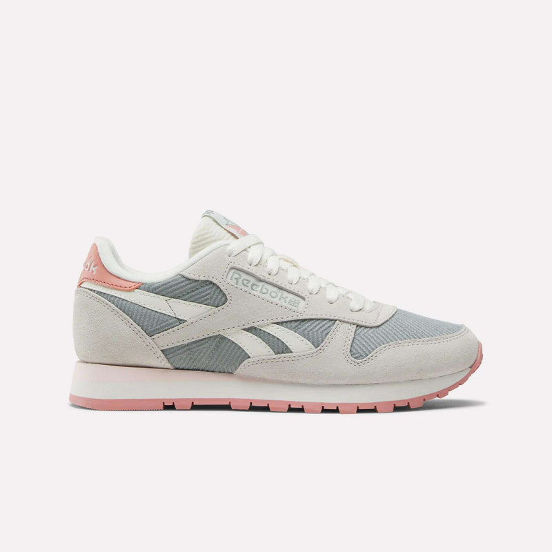 The Reebok Women's Classic Leather Shoes feature light grey and white panels, pale pink accents on the heel, outsole, and branding, suede details, a white lace-up closure, and a flat rubber sole for a retro look.