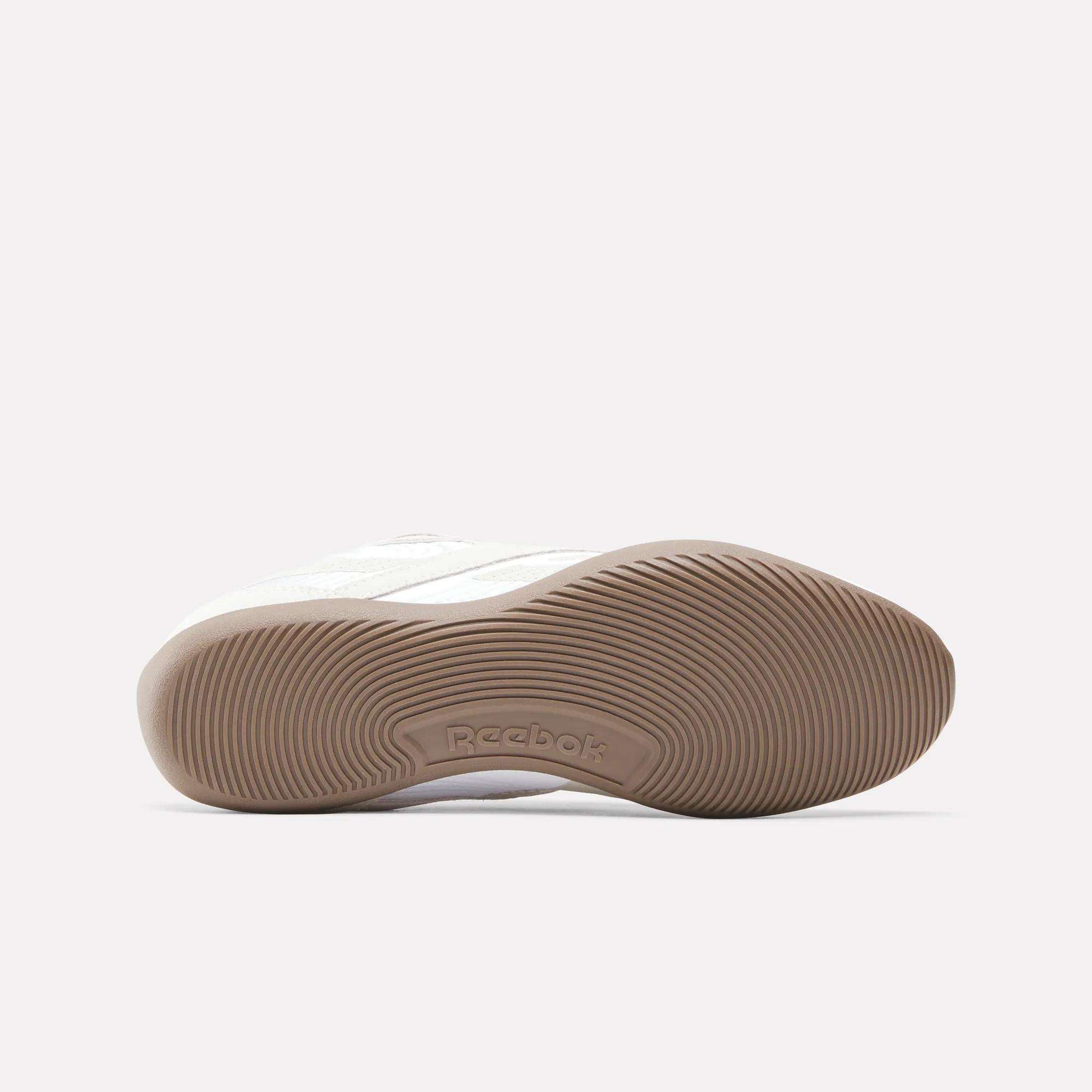 A close-up of the sole of a Reebok Women's Ultra Lo Shoe highlights the textured tan rubber outsole with curved grooves and the embossed Reebok logo at its center. The low-profile shoe is shown on its side against a plain background.