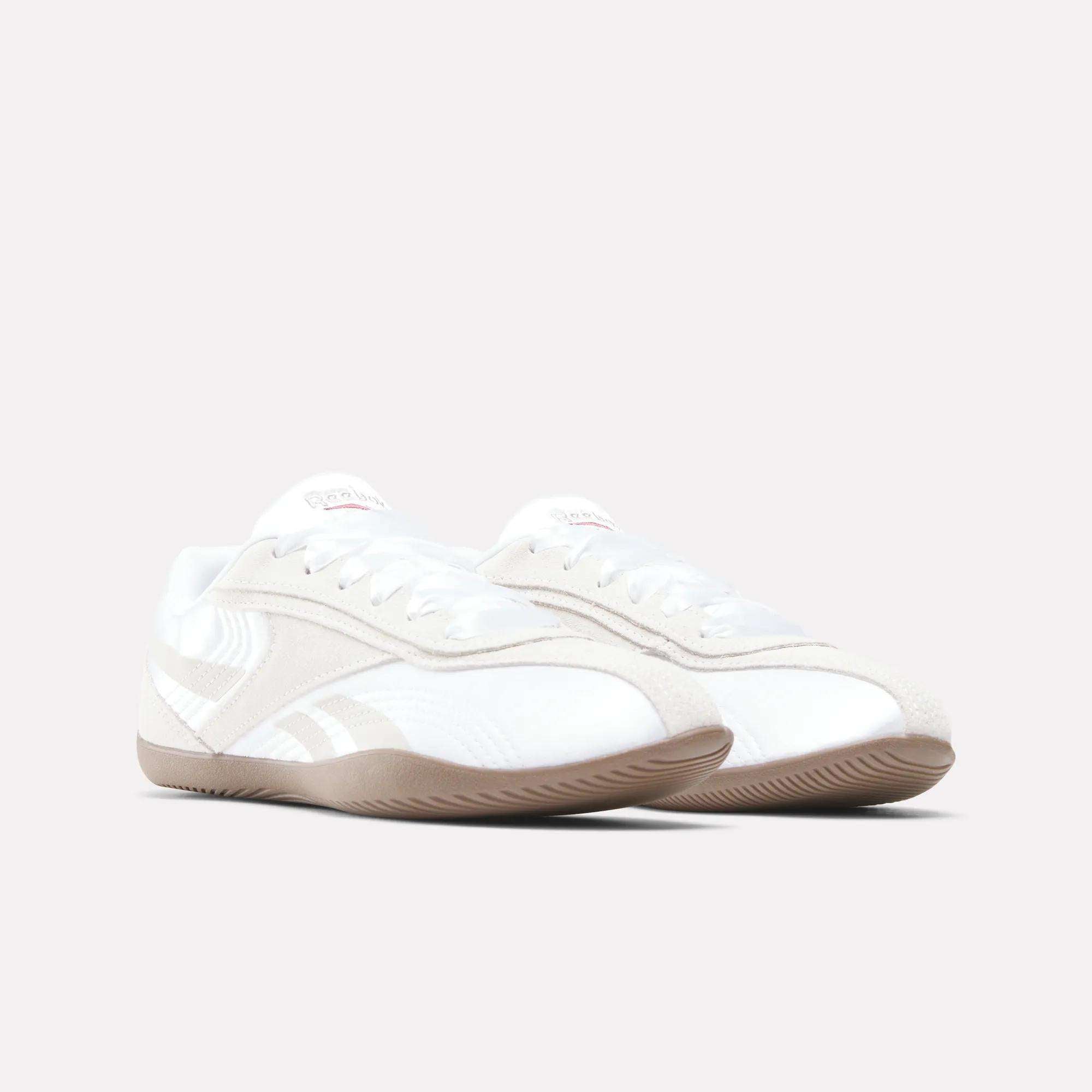 The Reebok Women's Ultra Lo Shoes feature a white and beige low-profile design with light brown rubber soles, white laces, and subtle side stripes, shown on a light gray background for a sport-inspired look.