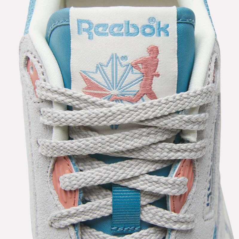 Close-up of the Reebok Women's Classic Nylon 89 Shoes tongue, showing the Reebok logo, a stylized maple leaf, and runner. This retro sneaker features white laces, off-white suede, light blue details, and light pink eyelets.