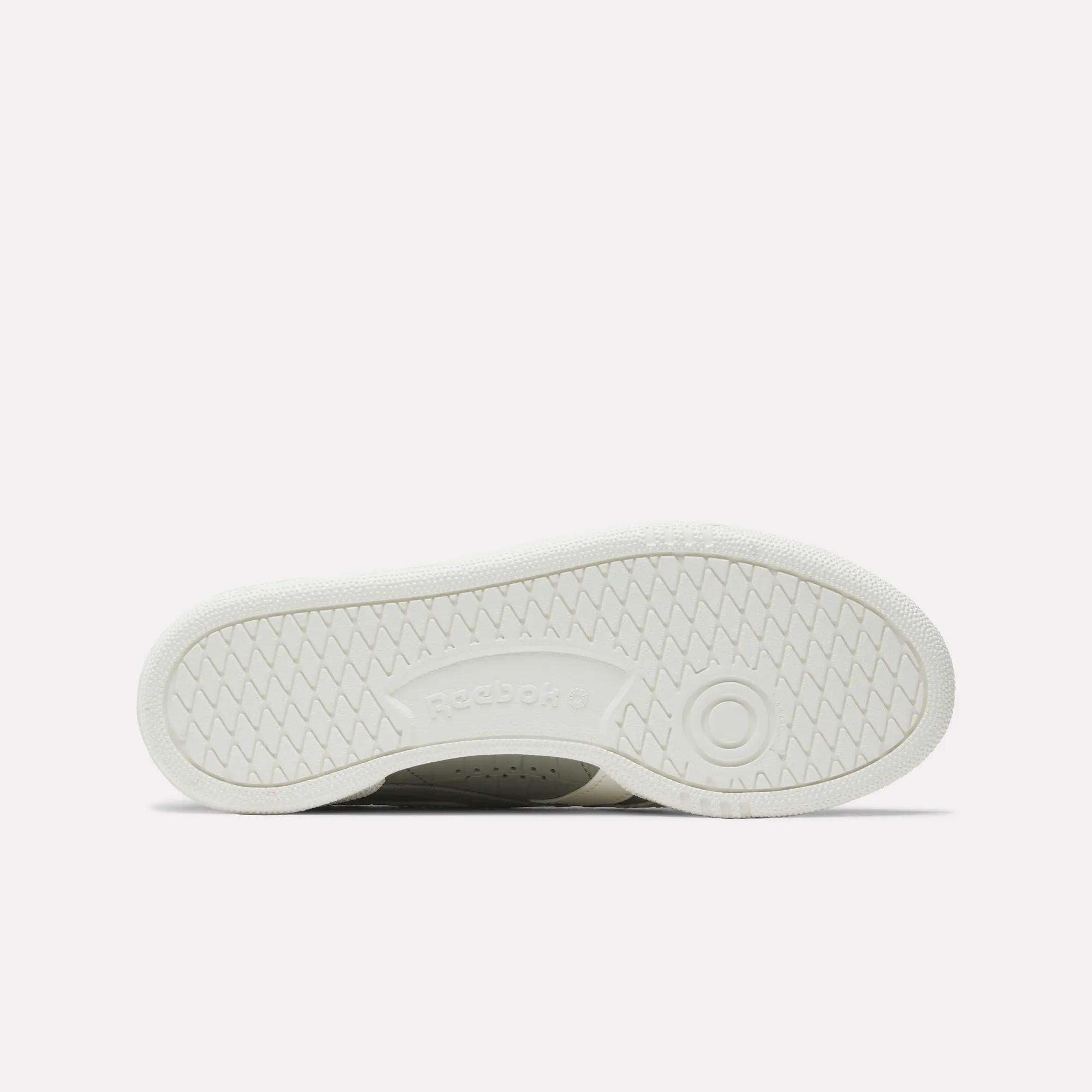 The image displays the white rubber outsole of Reebok Women's Club C Grounds UK Shoes, with a circular tread near the heel, herringbone traction patterns, and an embossed Reebok logo at the center.