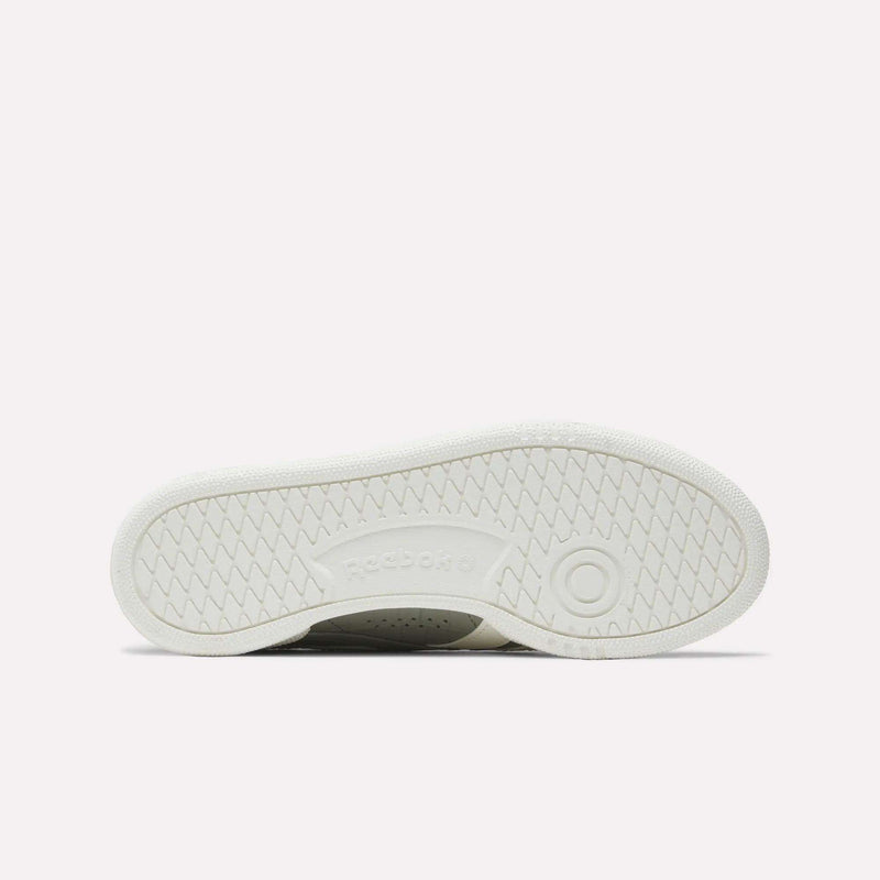 The image displays the white rubber outsole of Reebok Women's Club C Grounds UK Shoes, with a circular tread near the heel, herringbone traction patterns, and an embossed Reebok logo at the center.