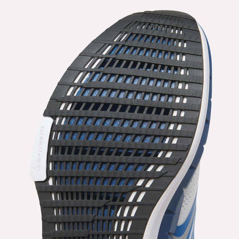Close-up of the Reebok Men's FloatZig Tread Running Shoes outsole, featuring a black, grooved tread for treadmill traction. Dual-density midsole and GRIP HONITEX on the white heel boost comfort; blue and white upper also shown.