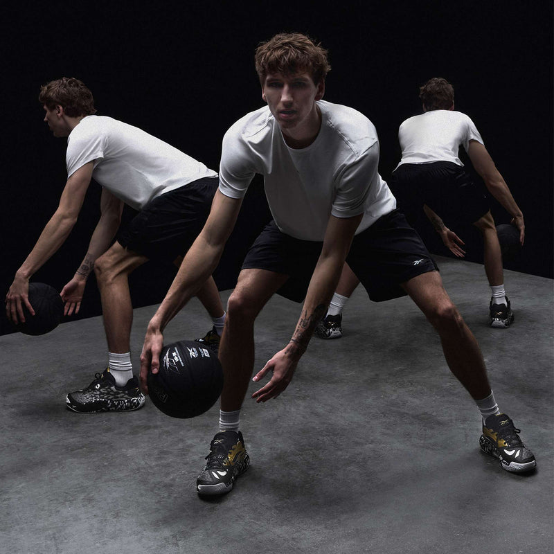 A man in a white t-shirt, black shorts, and Reebok Engine A 26 Basketball Shoes performs a dribbling move, shown in three overlapping positions to capture his motion on a gray floor against a dark background.
