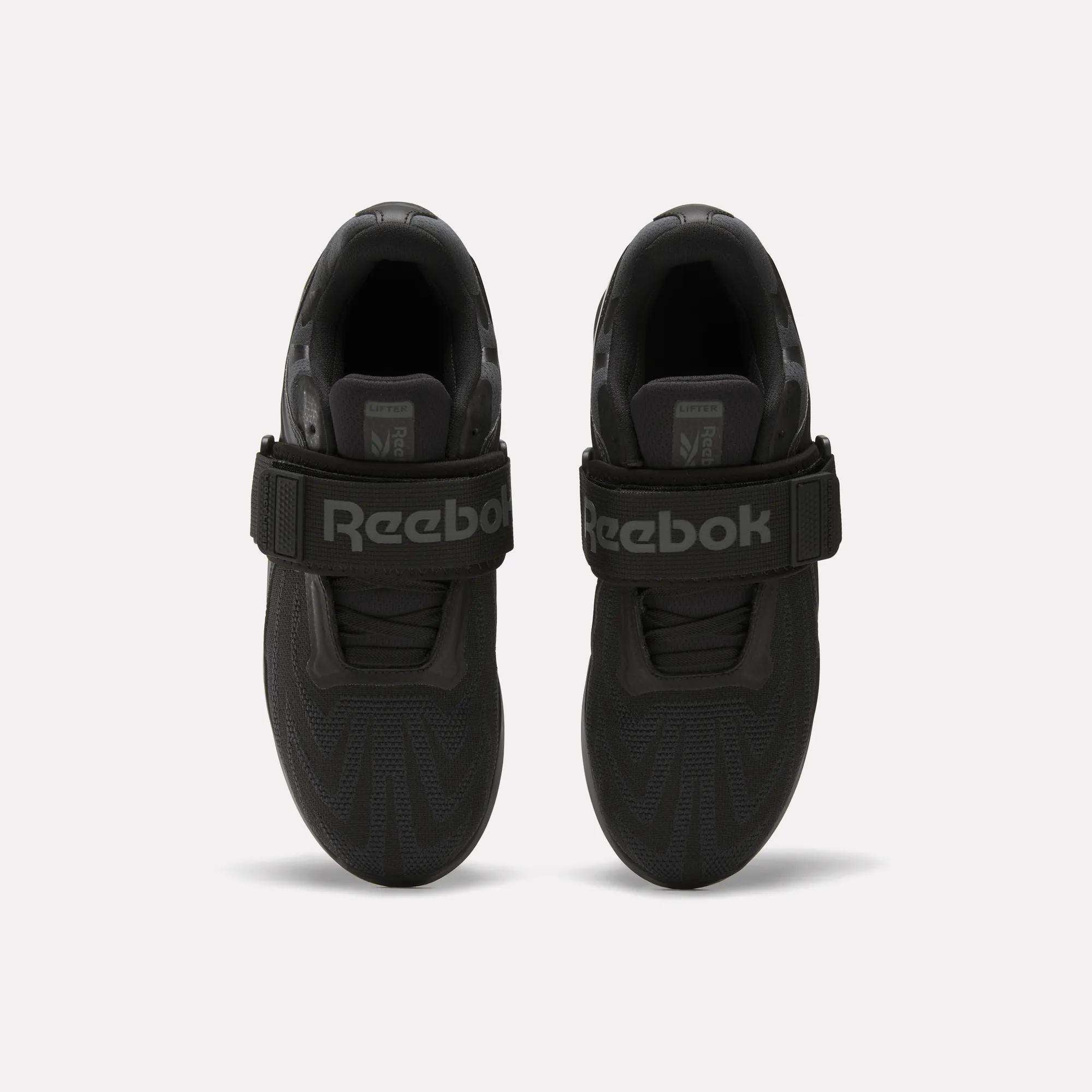Top view of a pair of Reebok Men's Legacy Lifter IV Shoes in black, with thick textured soles and wide velcro straps for stability. The large Reebok logo is visible. Set against a plain, light-colored background.