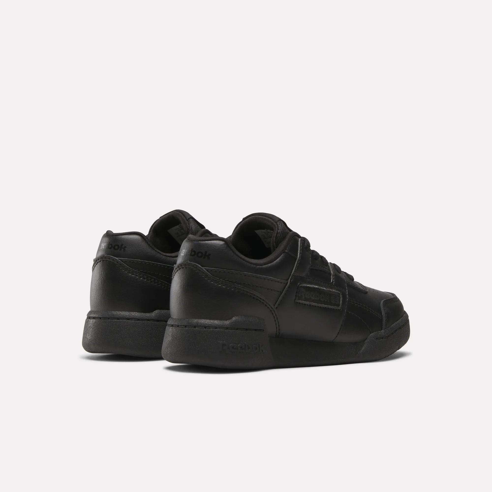 A pair of Reebok Workout Plus Shoes for little kids, shown in black from the back and side. These low-top sneakers feature laces and Reebok branding on the heel and side, displayed against a plain white background.