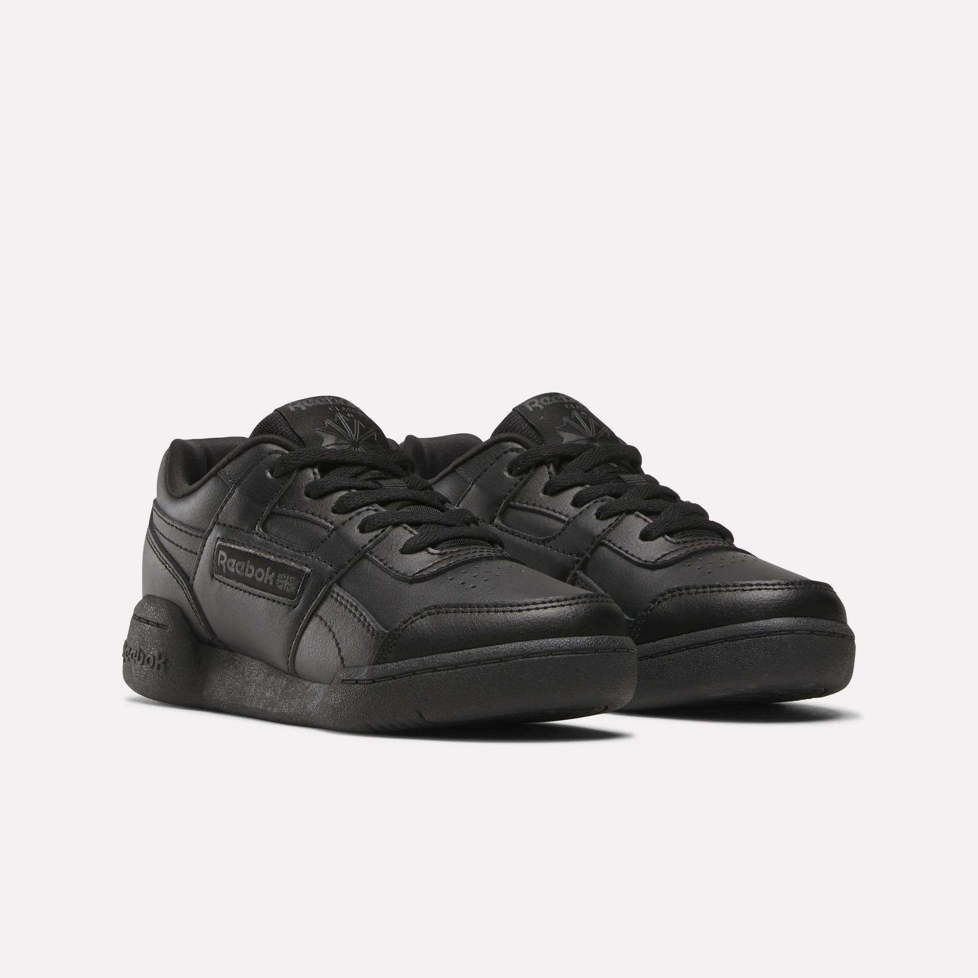 A pair of black Reebok Workout Plus Shoes for little kids, featuring a low-top design, lace-up fronts, and Reebok branding on the side and tongue, shown on a plain white background.