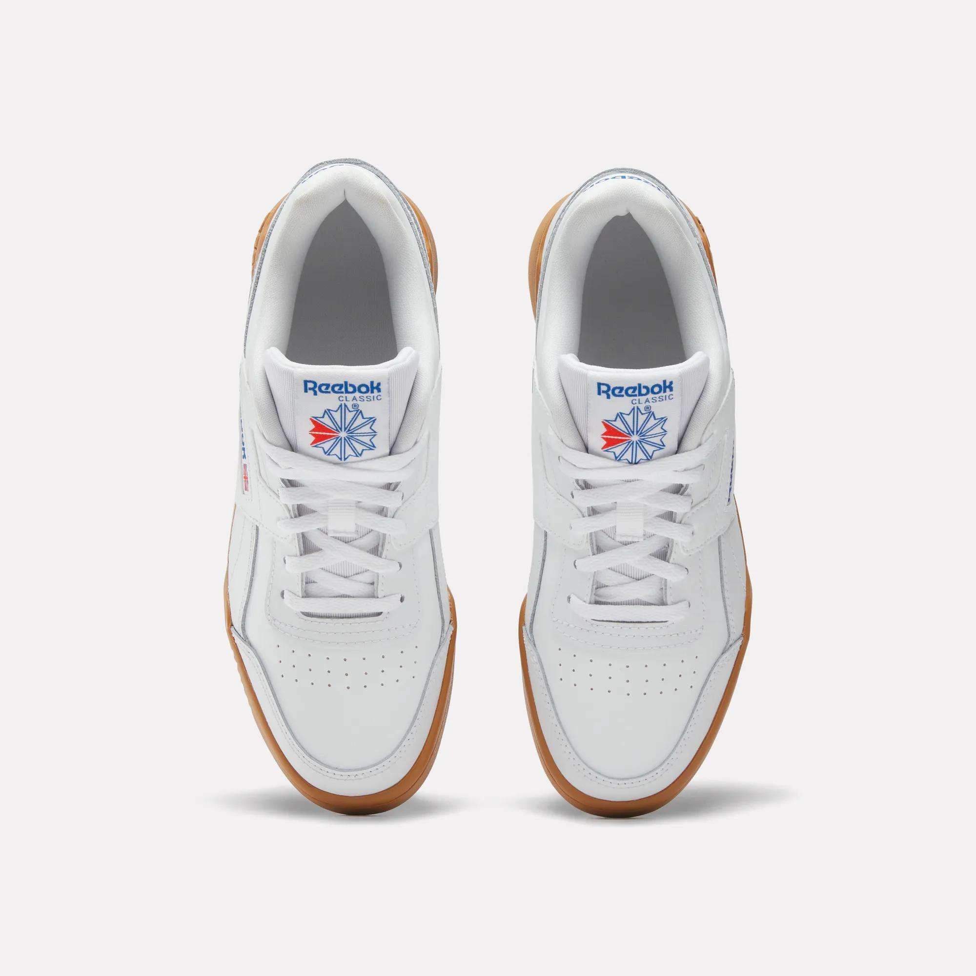 Top view of Reebok Workout Plus Shoes for big kids, featuring white retro uppers with gum soles, white laces, and blue and red Reebok logos on the tongues, placed side by side on a plain light background.