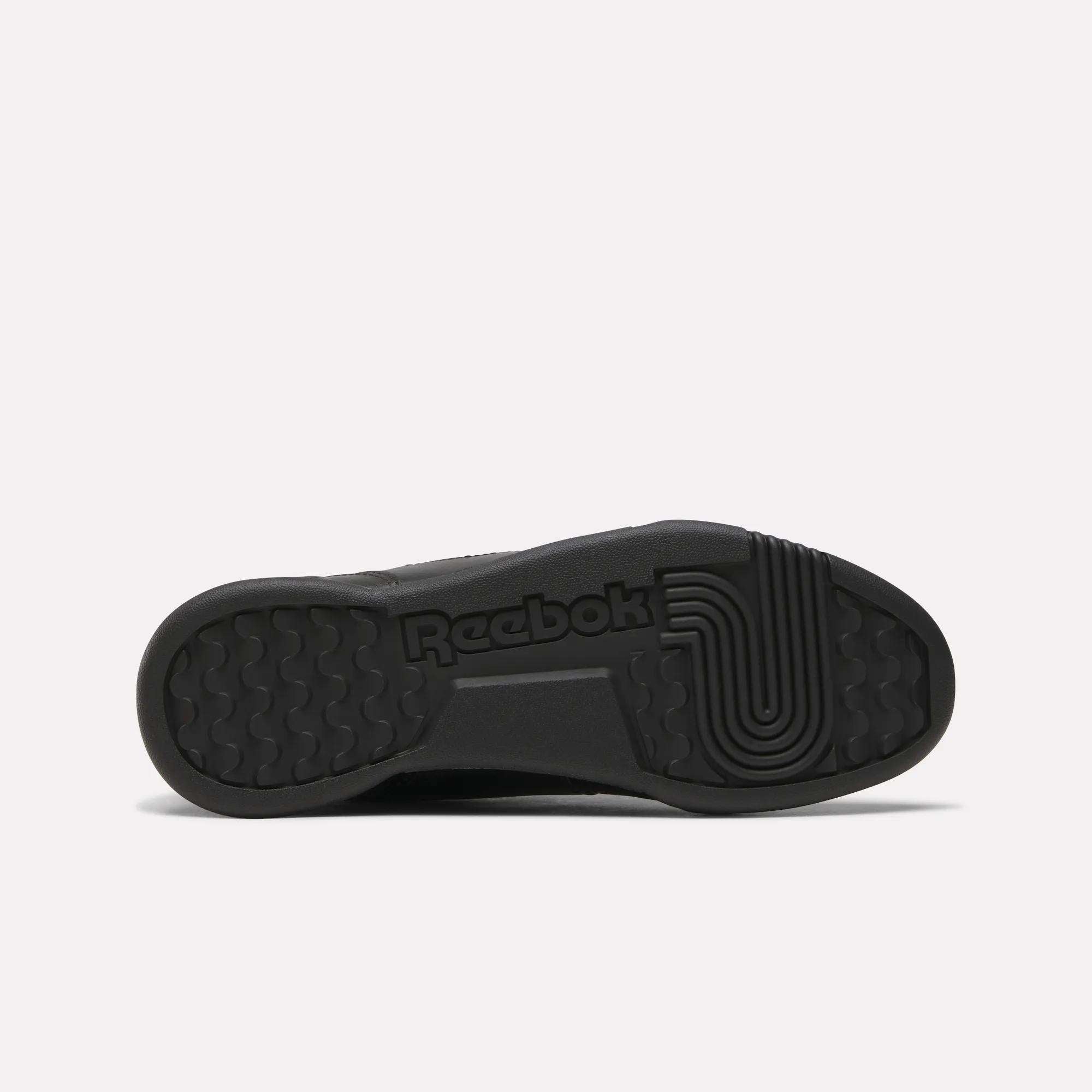 The image displays the black rubber sole of the Reebok Workout Plus Shoes for Big Kids, featuring the Reebok logo and circular tread for grip. The sole blends geometric and textured patterns, emphasizing these sporty sneakers' authentic style.