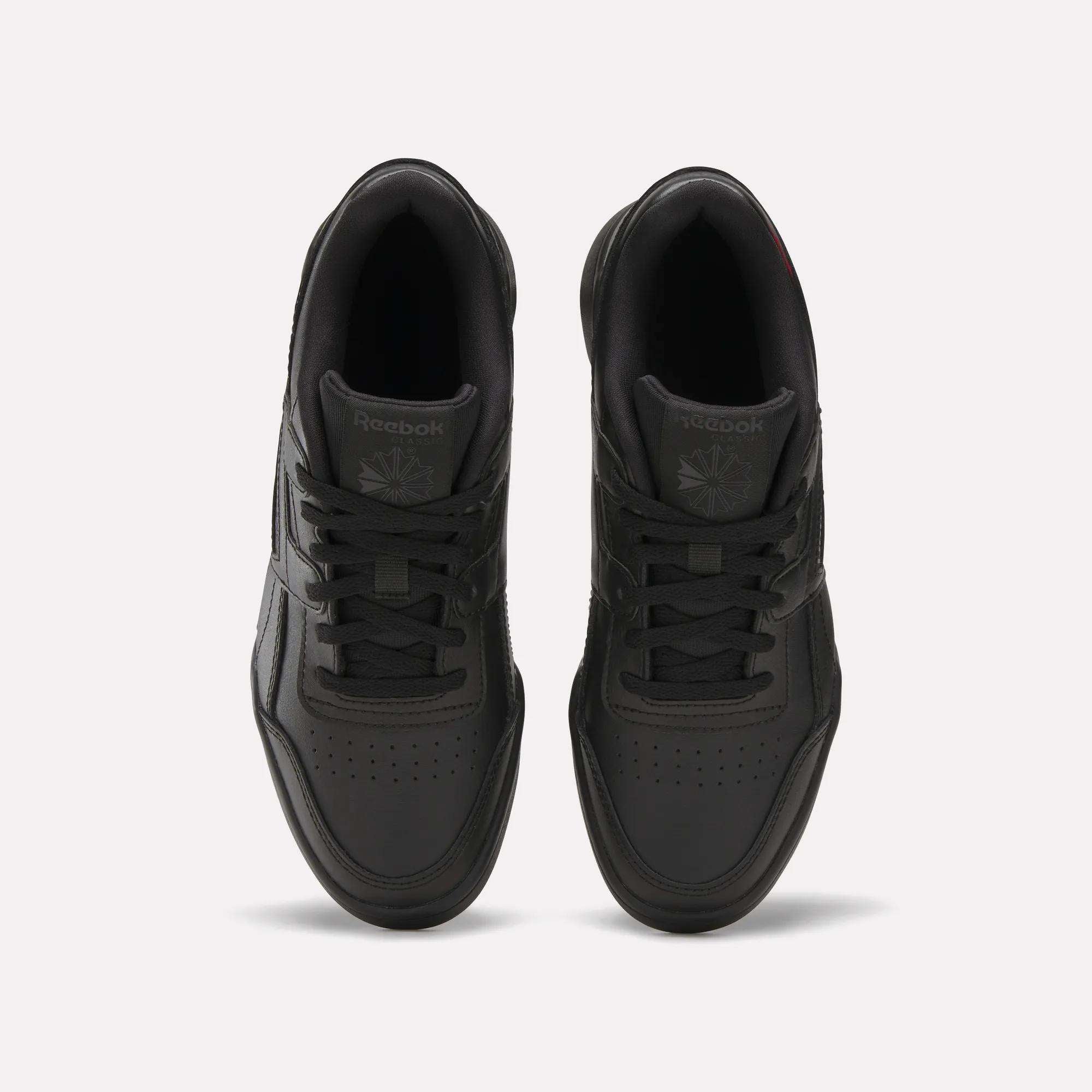 Top view of a pair of black Reebok Workout Plus Shoes for big kids with laces, displayed side by side on a white background. The Reebok logo appears on the tongue of each sneaker.