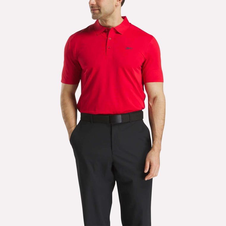 A man models the Reebok Men's Short Sleeve Bonded Micro Pique Polo Shirt in bright red and black pants against a white background, his left hand in his pocket to highlight the shirt's moisture-wicking comfort.