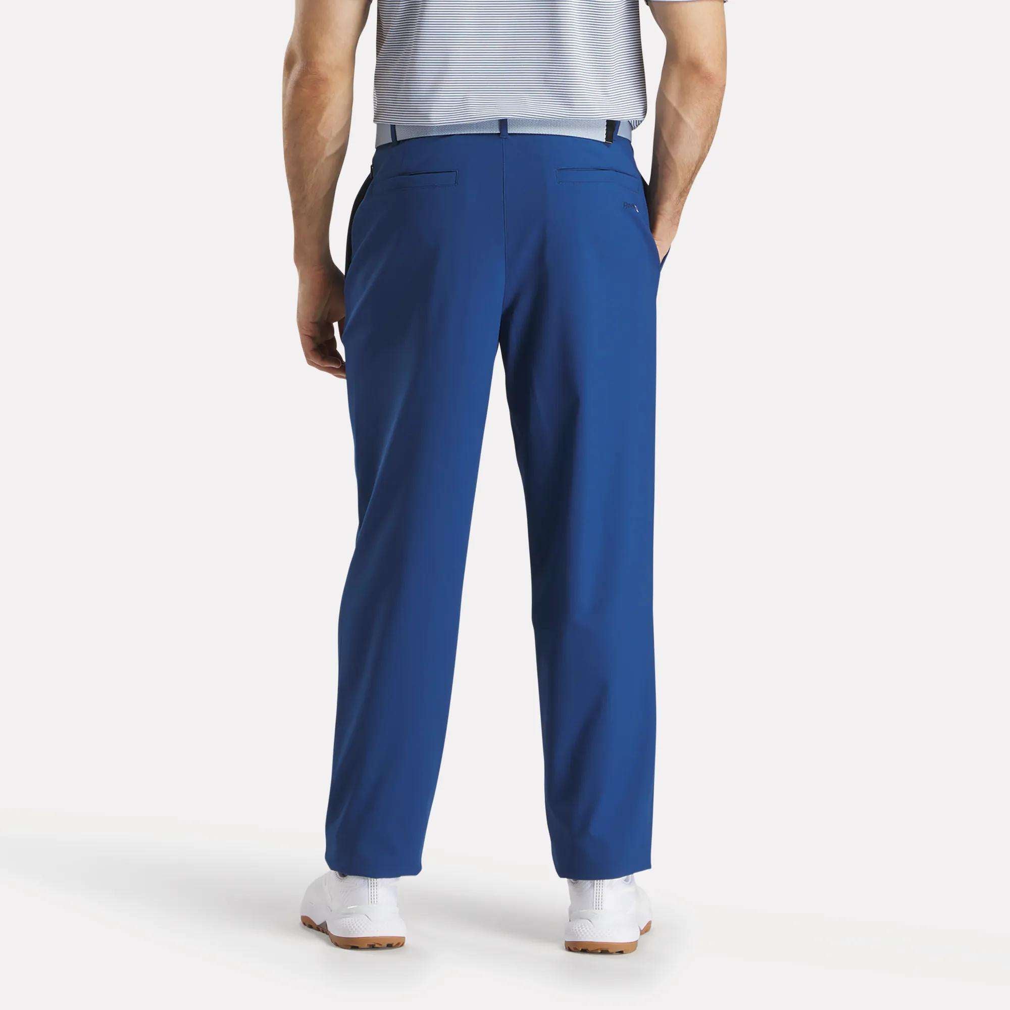 A man faces away in Reebok Men's The Favorite Golf Pants - Relaxed Fit (bright blue), paired with a tucked-in light gray striped shirt and white sneakers. His hands are in his pockets against a plain white background.