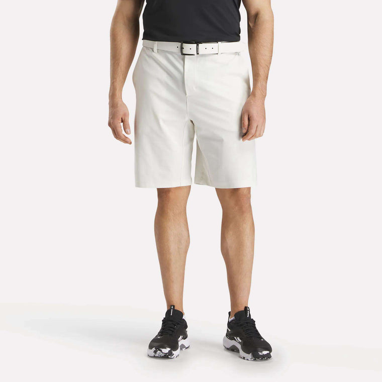 A man stands facing forward against a plain white background wearing a black shirt, white belt, Reebok Men's The Favorite Golf Shorts - 9" Inseam in white, and black-and-white sneakers.