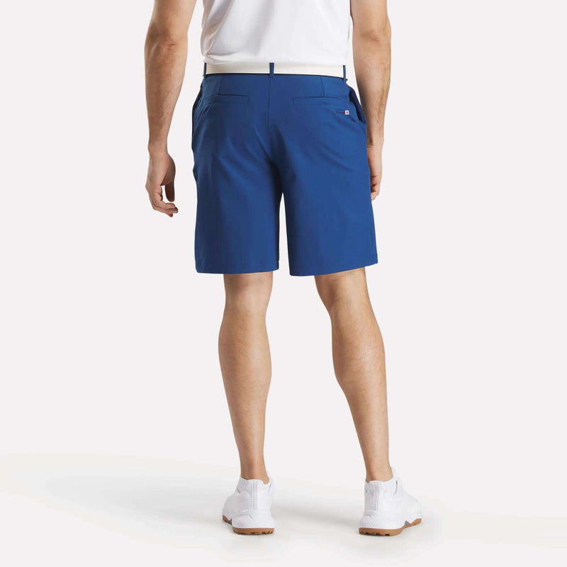A man stands facing away, wearing Reebok Men's The Favorite Golf Shorts - 9" Inseam made with sweat-wicking four-way stretch fabric, paired with a white polo shirt, white belt, and white sneakers against a plain white background.