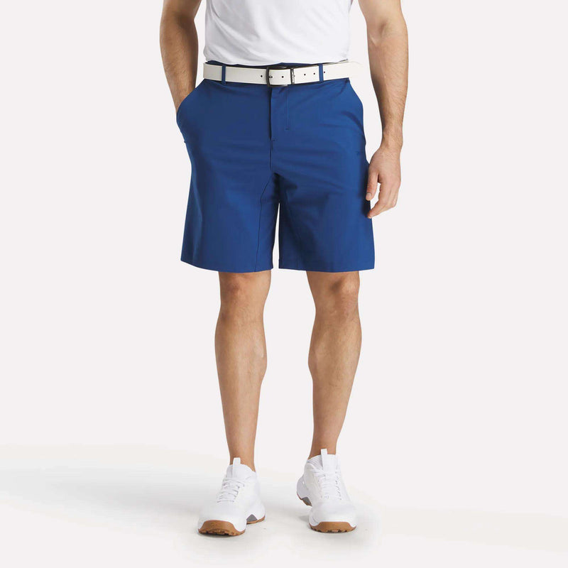 A man stands with hands in the pockets of his Reebok Men's The Favorite Golf Shorts - 9" Inseam, made from four-way stretch fabric. He wears a white belt, T-shirt, and athletic shoes. The photo is cropped at the shoulders; his face is not visible.
