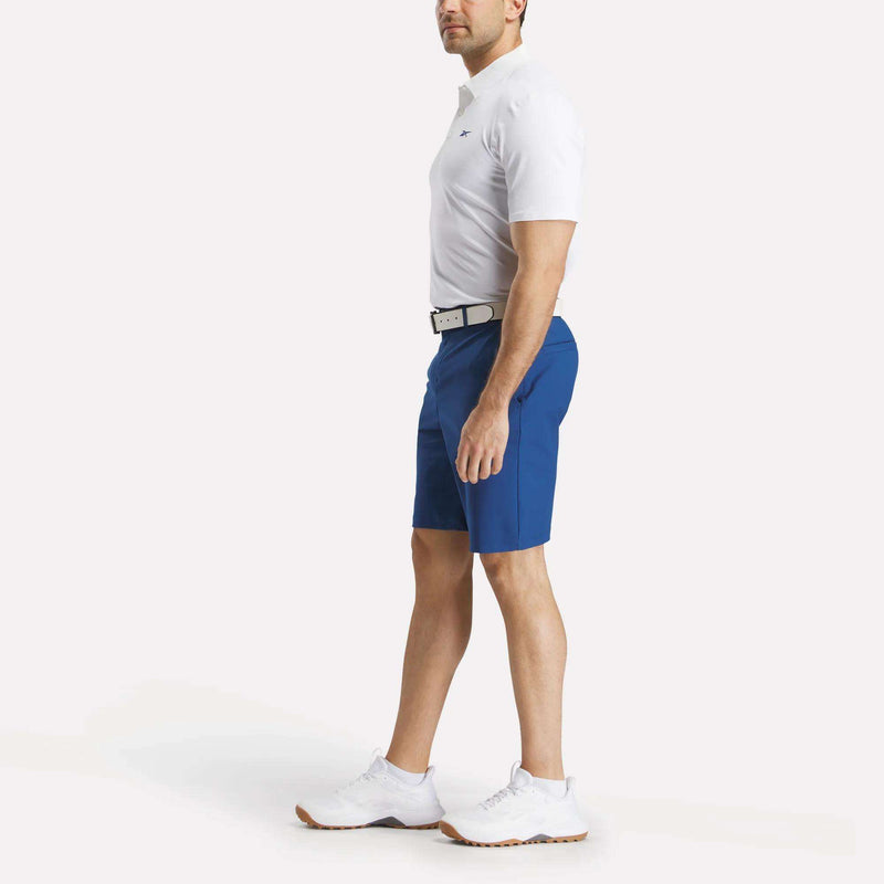 A man stands sideways against a plain light background, wearing a white polo shirt, white belt, white athletic shoes, and Reebok Men's The Favorite Golf Shorts - 9" Inseam in blue made of four-way stretch fabric.