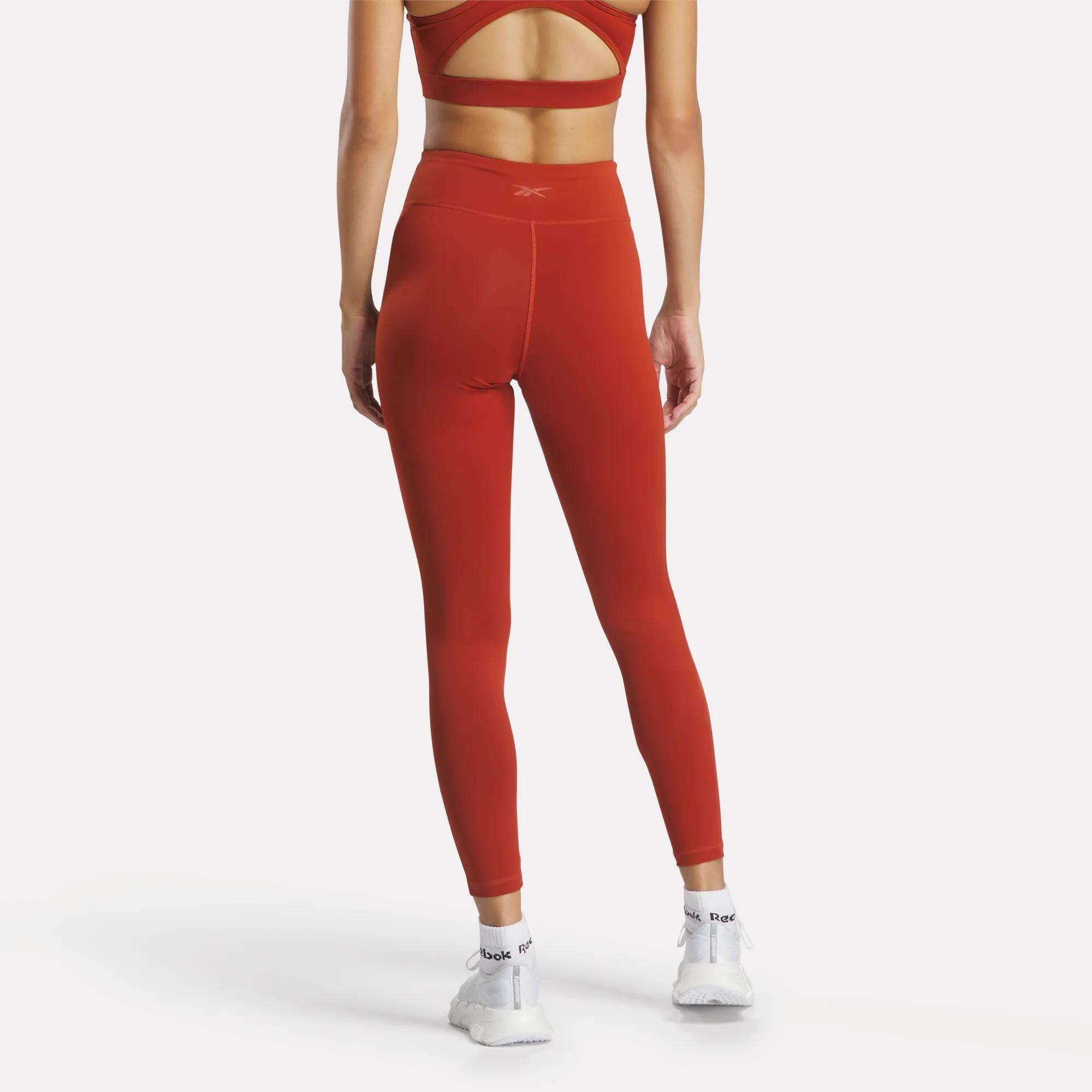 A woman stands with her back to the camera, wearing Reebok Women's Lux High-Rise Tights and a matching sports bra in rust, made from RBK-DRY performance fabric, paired with white athletic shoes. The background is plain white.