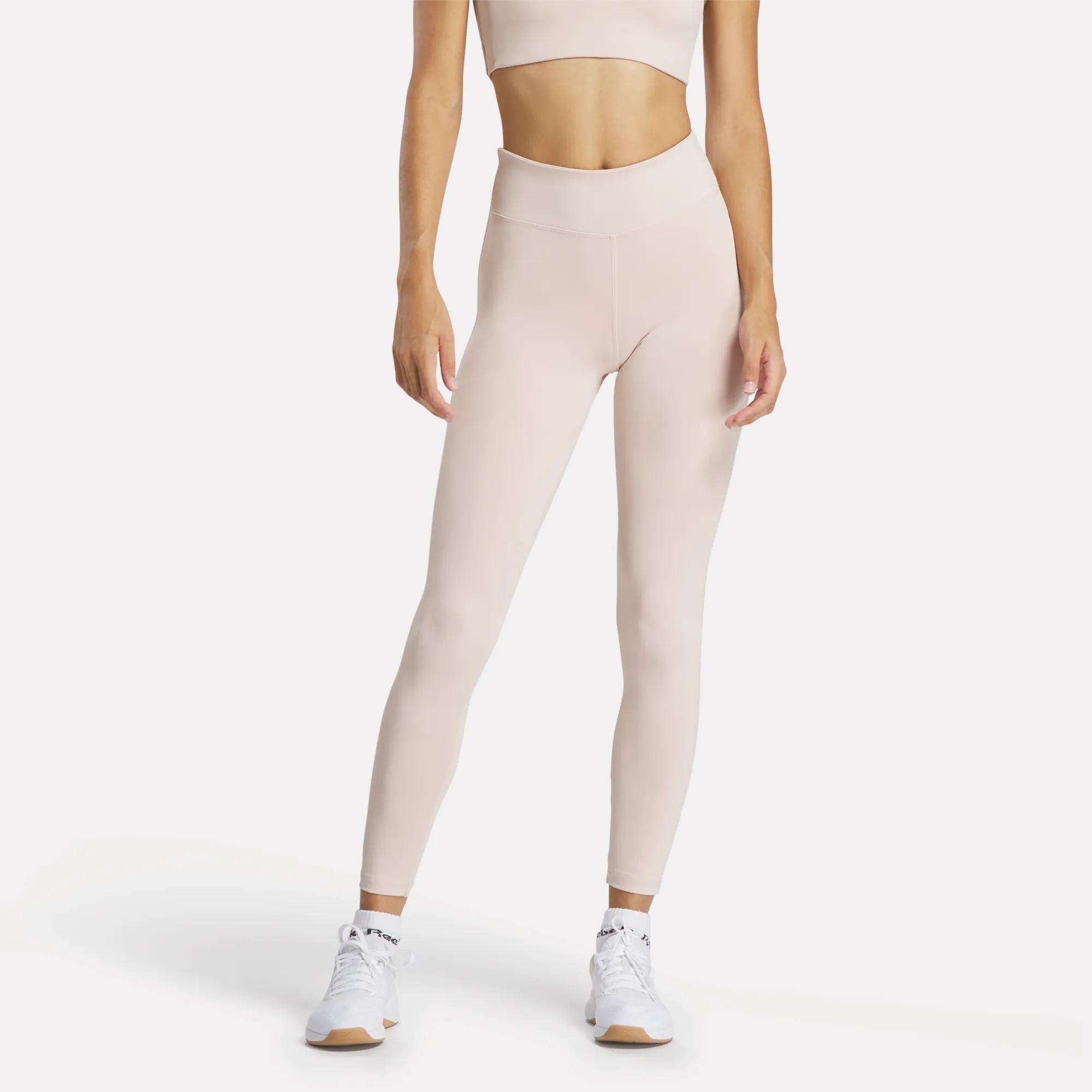 A woman models Reebok Women's Lux High-Rise Tights with a light beige sports bra and white sneakers, standing relaxed against a plain background. The RBK-DRY fabric keeps her comfortable and stylish.