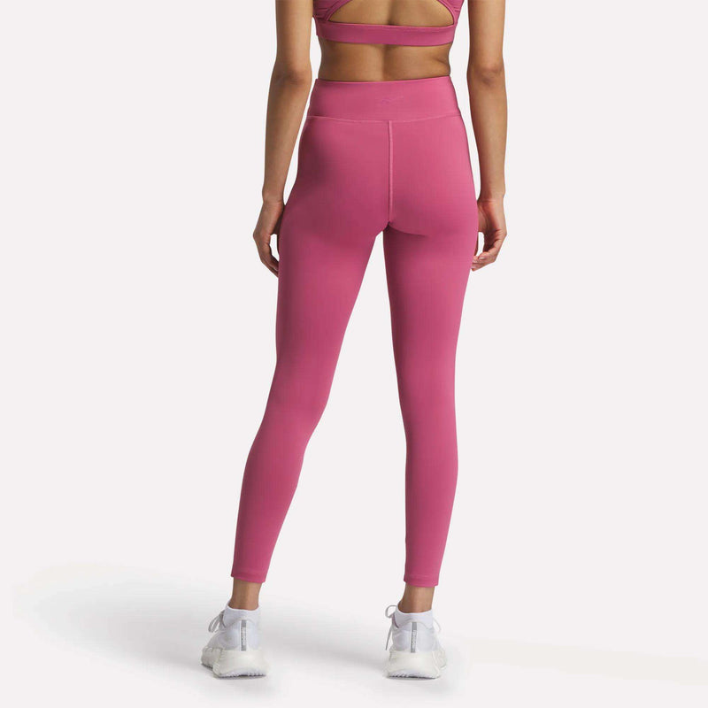 A woman faces away from the camera wearing Reebok Women's Lux-Lite High-Rise Tights with a matching pink sports bra, both made with RBK-DRY fabric, and white athletic shoes against a plain white background.