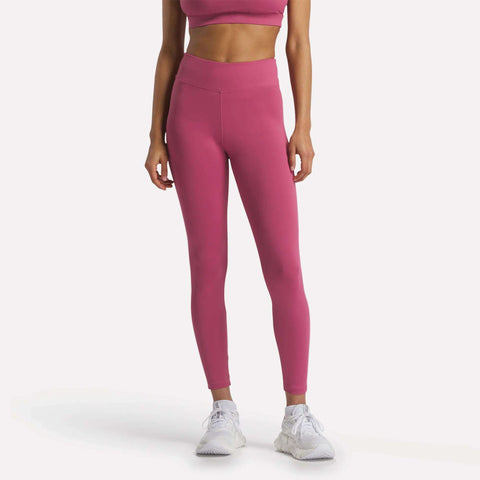 A woman models Reebok Women’s Lux-Lite High-Rise Tights and a pink sports bra with RBK-DRY fabric, paired with white sneakers, against a plain white background. Her head and lower legs are out of frame.