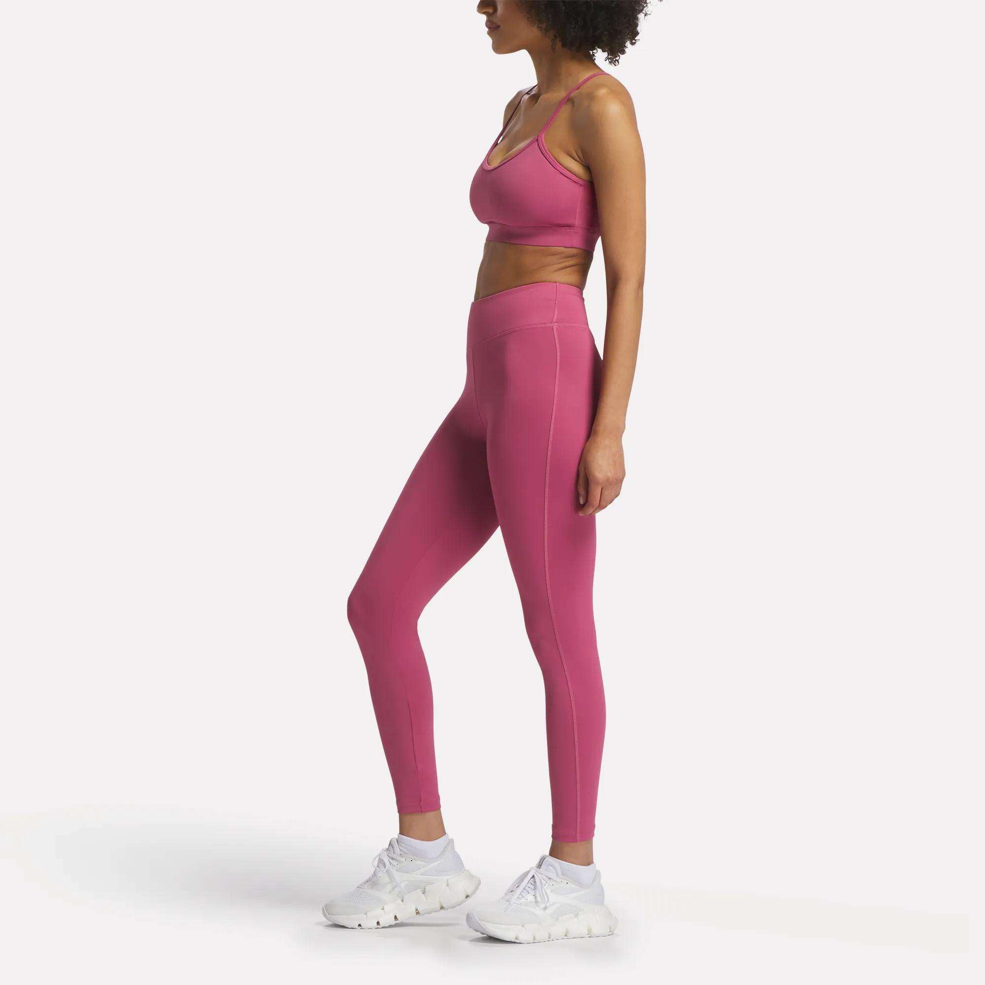 A woman stands in profile wearing Reebok Women's Lux-Lite High-Rise Tights and a matching pink sports bra with white sneakers, set against a plain white background.