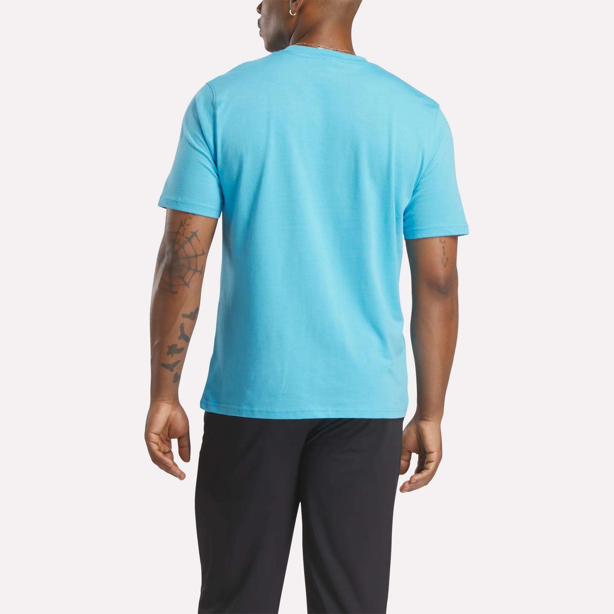 A man faces away, wearing a bright blue Reebok Men's Big Logo Crewneck Short Sleeve Tee and black pants, displaying the shirt's back. His arms hang relaxed at his sides, showing tattoos on his left arm.