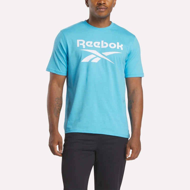 A man stands against a plain light background, wearing Reebok's Men's Big Logo Crewneck Short Sleeve Tee in light blue with a large white logo, paired with black pants.
