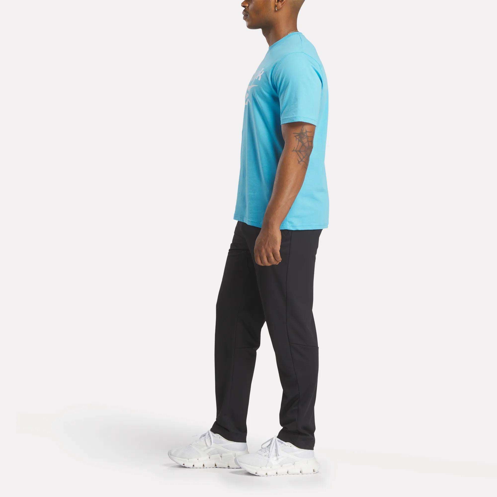 A man in profile poses against a plain white background wearing the Reebok Men's Big Logo Crewneck Short Sleeve Tee in light blue, black pants, and white sneakers. He has a tattoo on his left arm.