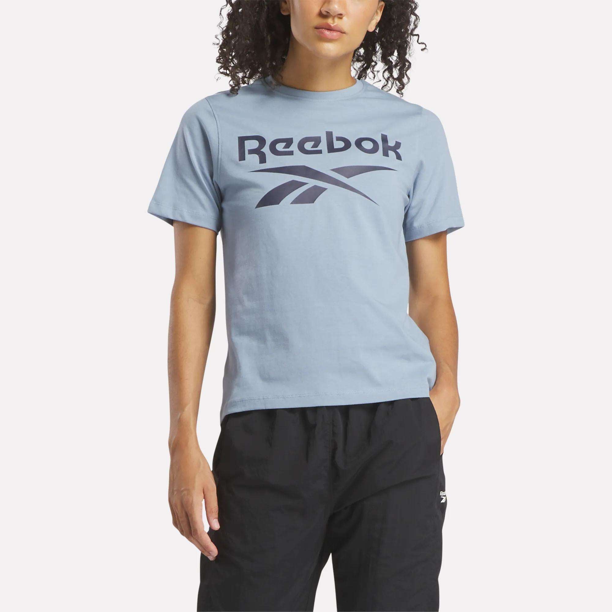 A person stands against a plain, light background wearing the Reebok Women's Big Logo Crewneck Short Sleeve Tee in light blue, made of soft cotton jersey with a large navy logo, paired with black pants.