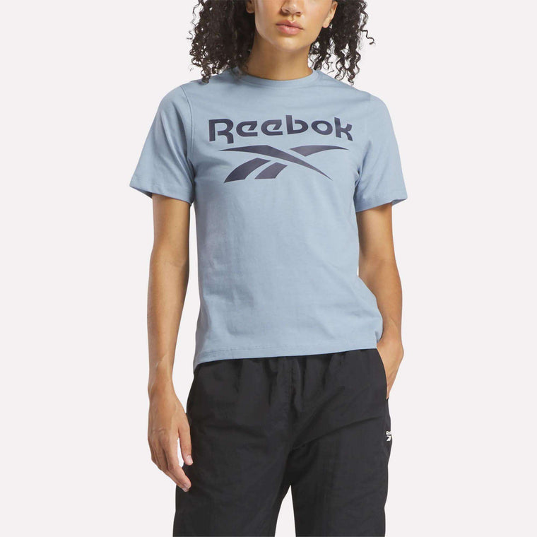 A person stands against a plain, light background wearing the Reebok Women's Big Logo Crewneck Short Sleeve Tee in light blue, made of soft cotton jersey with a large navy logo, paired with black pants.