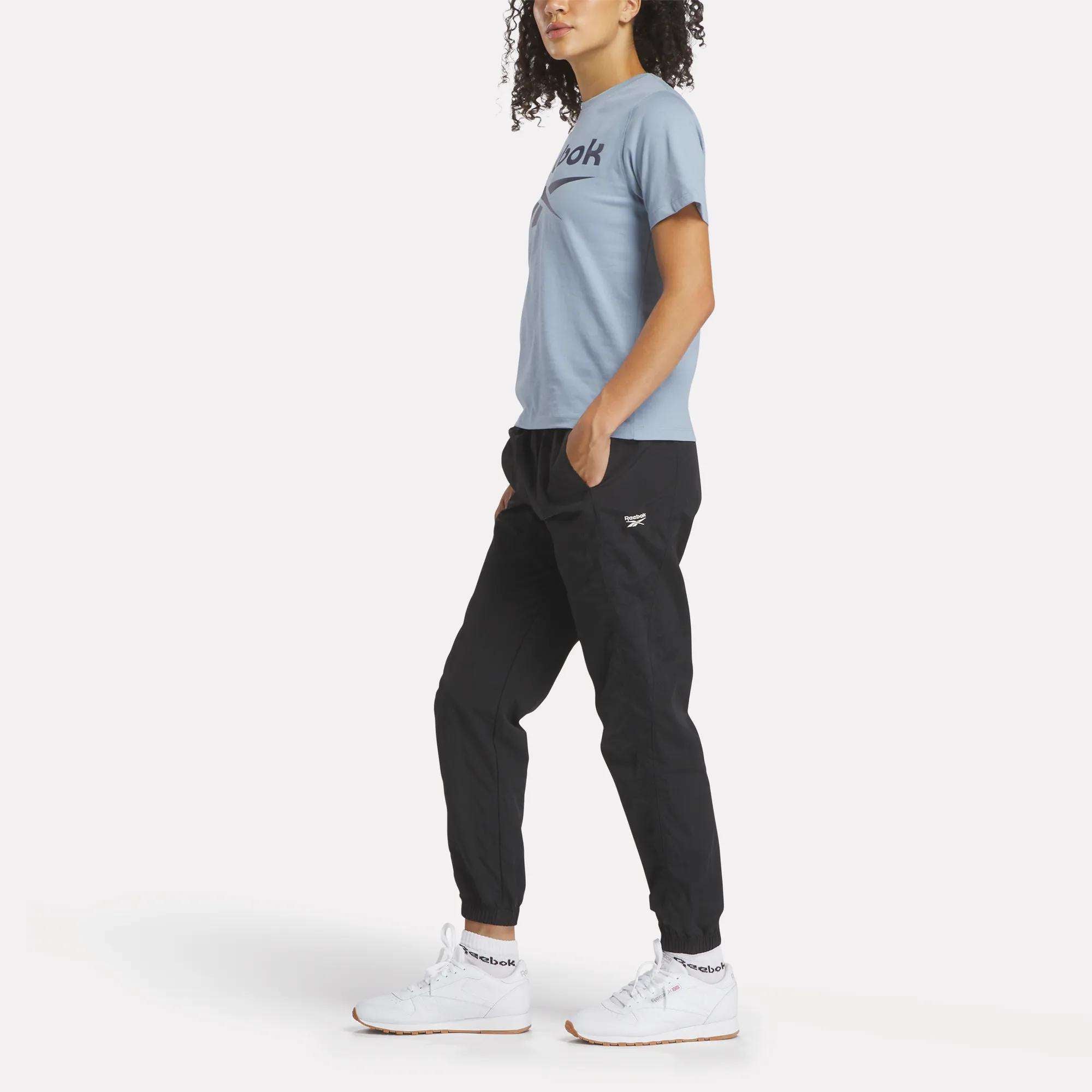 A person with curly hair stands sideways, wearing a light blue Reebok Women's Big Logo Crewneck Short Sleeve Tee, black jogger pants, and white Reebok sneakers against a plain white background.