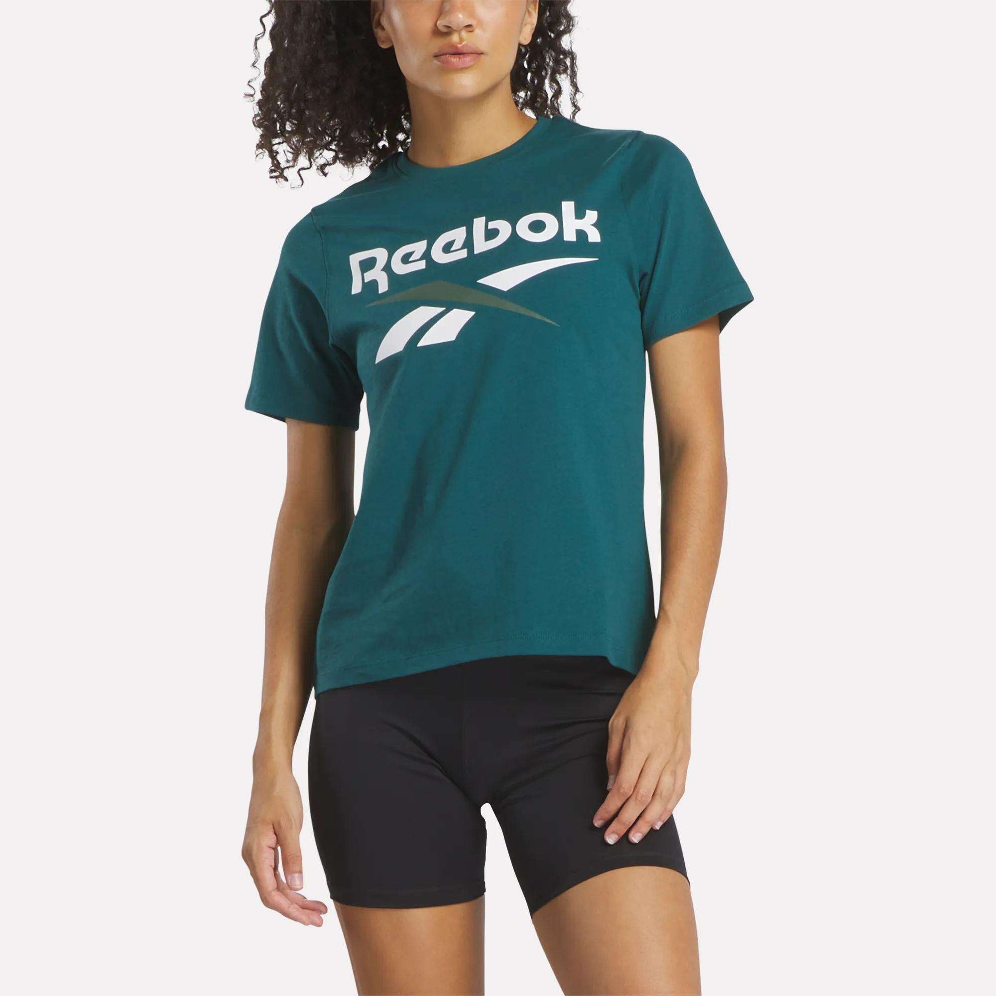 A woman wearing the Reebok Women's Two-Color Logo Crew Short Sleeve Tee stands against a light background, paired with black shorts.