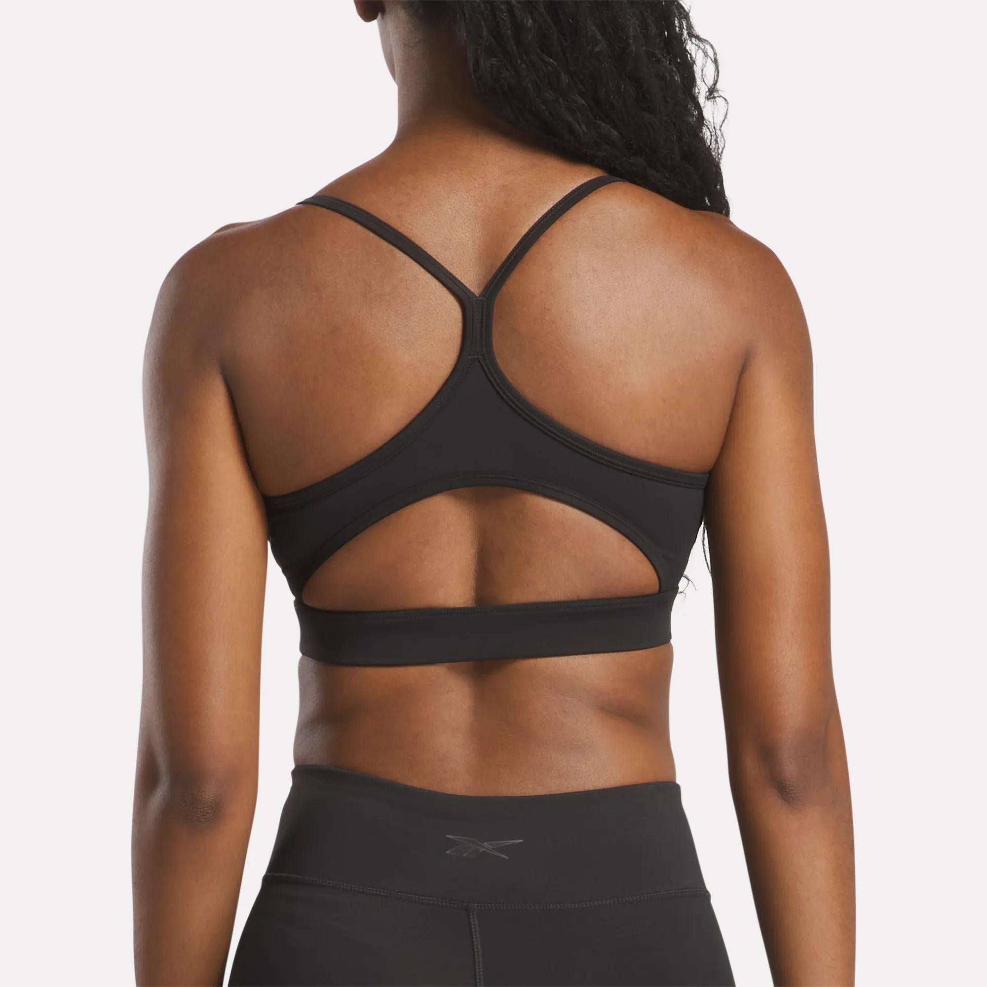 A woman wears the Reebok Women's Yoga Strappy Bra, featuring crisscross straps and a cutout back for medium support, paired with high-waisted black leggings and shown from behind against a plain background.